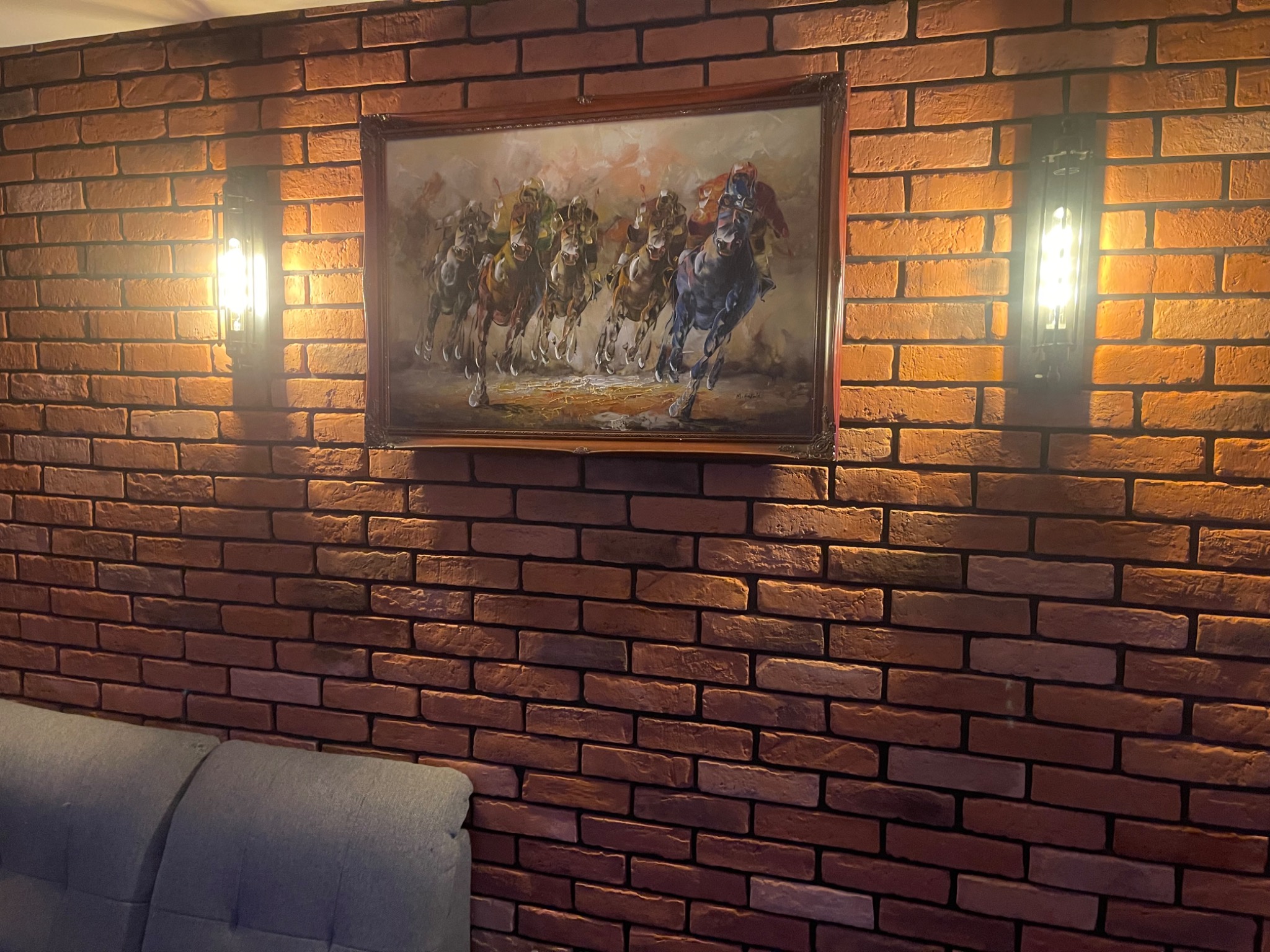 Ceglana ściana z obrazem koni i dwoma kinkietami po bokach, oświetlającymi dekoracyjną powierzchnię. Fragment szarej sofy w dolnej części kadru.