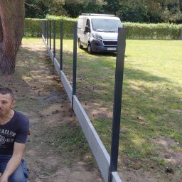 Art-bau usługi budowlane - Montaż prefabrykowanego ogrodzenia betonowego z metalowymi słupkami na trawiastym terenie. W tle biały bus i zielony żywopłot.