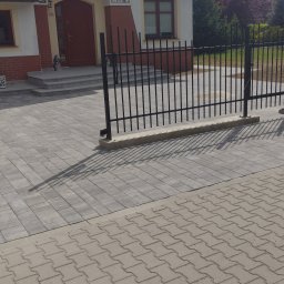 Art-bau usługi budowlane - Dwukondygnacyjny dom z balkonem, widoczny zza czarnego ogrodzenia i szarej kostki brukowej. Tabliczka z numerem 'Cisowa 12' nad wejściem.