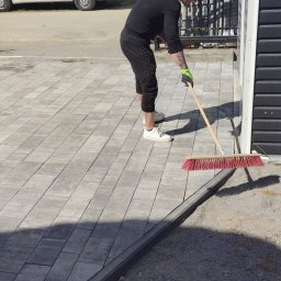 Art-bau usługi budowlane - Układanie kostki brukowej w Prochowicach: mężczyzna zamiata nową nawierzchnię z szarej kostki, widoczny fragment domu i samochód dostawczy.