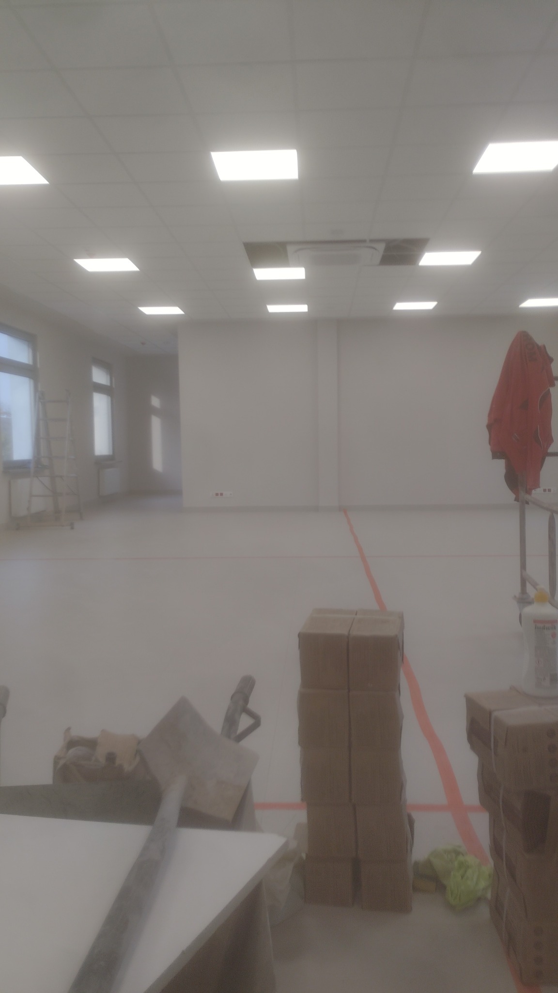 Wnętrze w trakcie prac wykończeniowych: widoczne kartony z materiałami, narzędzia, drabina i wykończona podłoga w dużym pomieszczeniu z oknami i równomiernym oświetleniem.