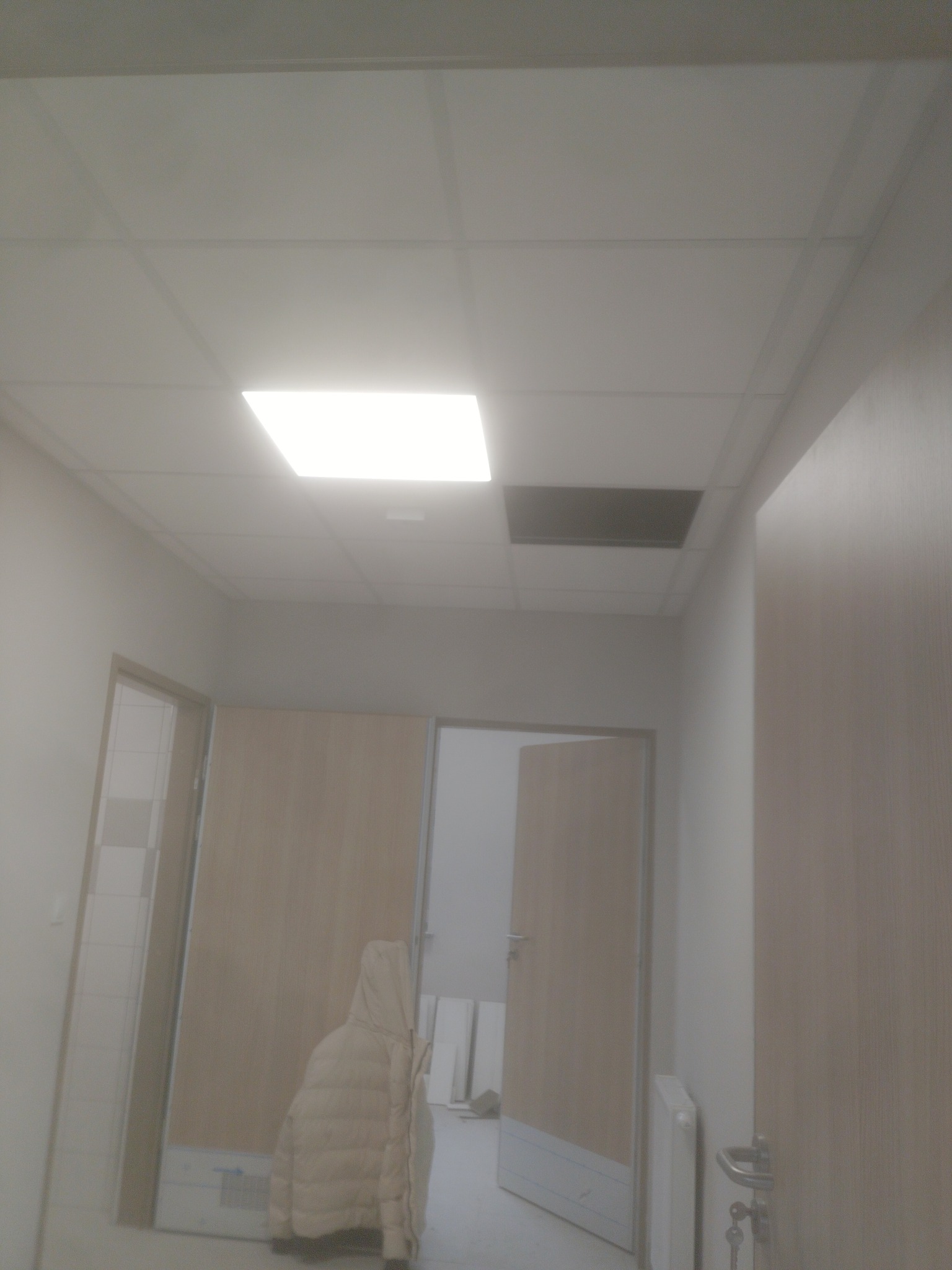 Korytarz z podwieszanym sufitem, gdzie brakuje jednej płyty. Widoczne drzwi i fragment grzejnika. Ujęcie z dołu, oświetlenie sufitowe.