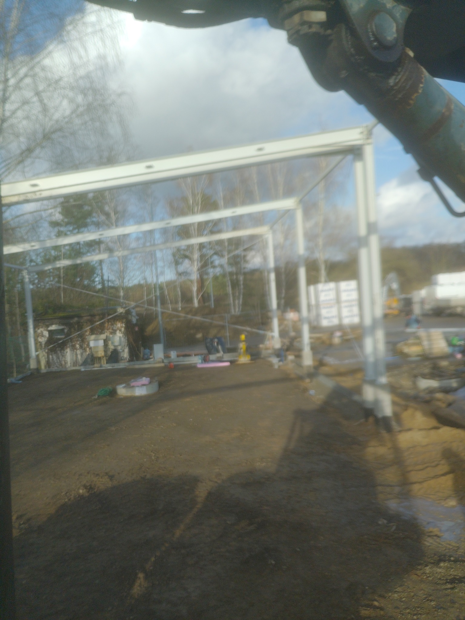 Stalowa konstrukcja szkieletowa budynku w trakcie montażu, widok z perspektywy operatora maszyny budowlanej, w tle drzewa i materiały budowlane.