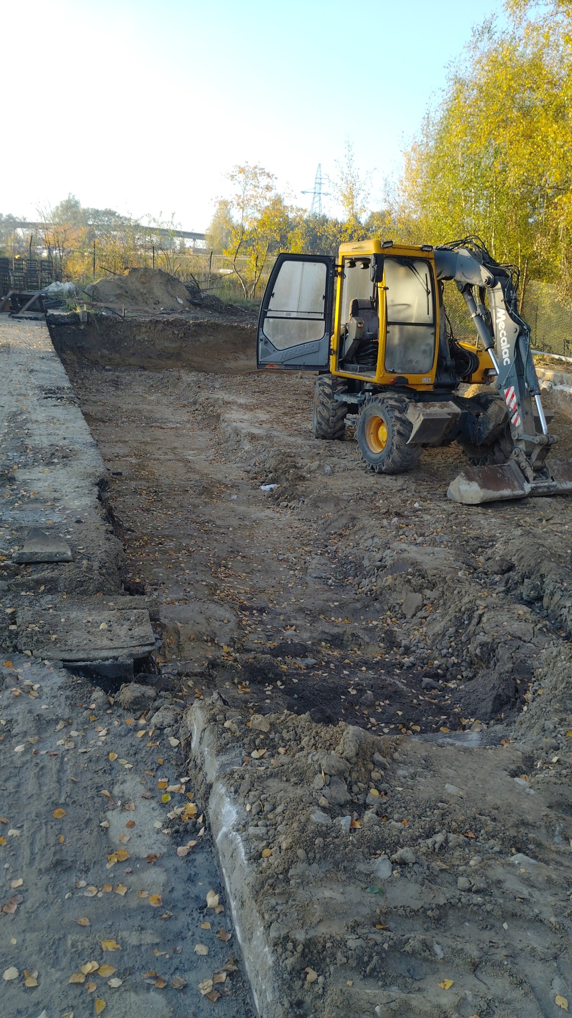 Żółta koparka Mecalac na placu budowy, z otwartymi drzwiami kabiny, przygotowuje teren pod fundamenty. Widoczne wykopane rowy i jesienne drzewa w tle.