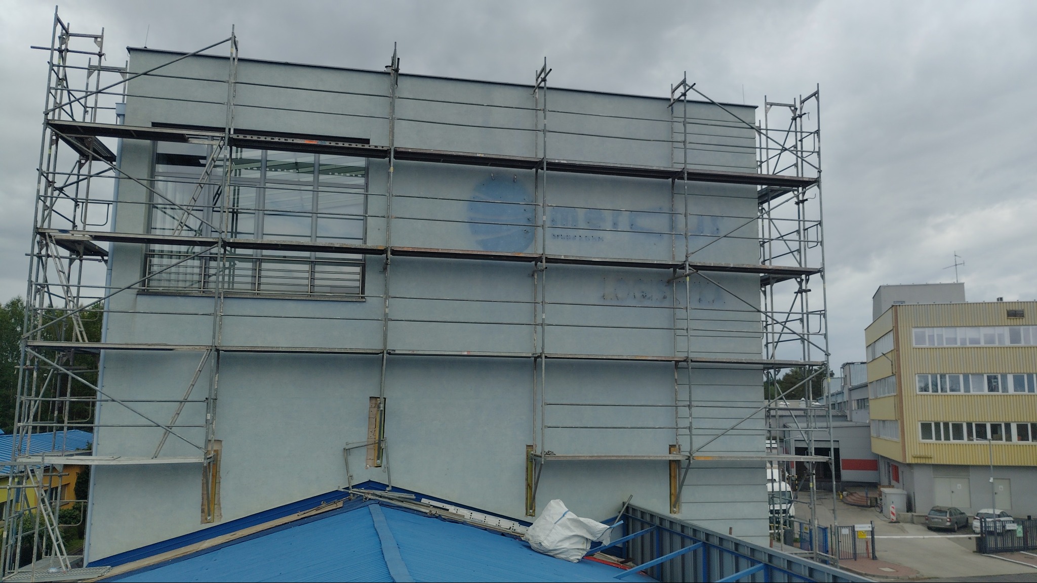 Elewacja budynku w trakcie prac, otoczona rusztowaniami. Widoczne ślady po usuniętym logo na ścianie. Niebo pochmurne, w tle budynki.