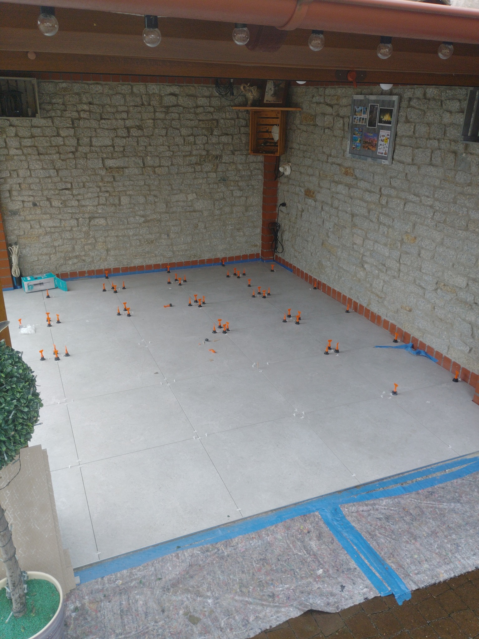 Układanie jasnych płytek podłogowych z użyciem pomarańczowych klipsów poziomujących w pomieszczeniu z kamiennymi ścianami i oświetleniem sufitowym.