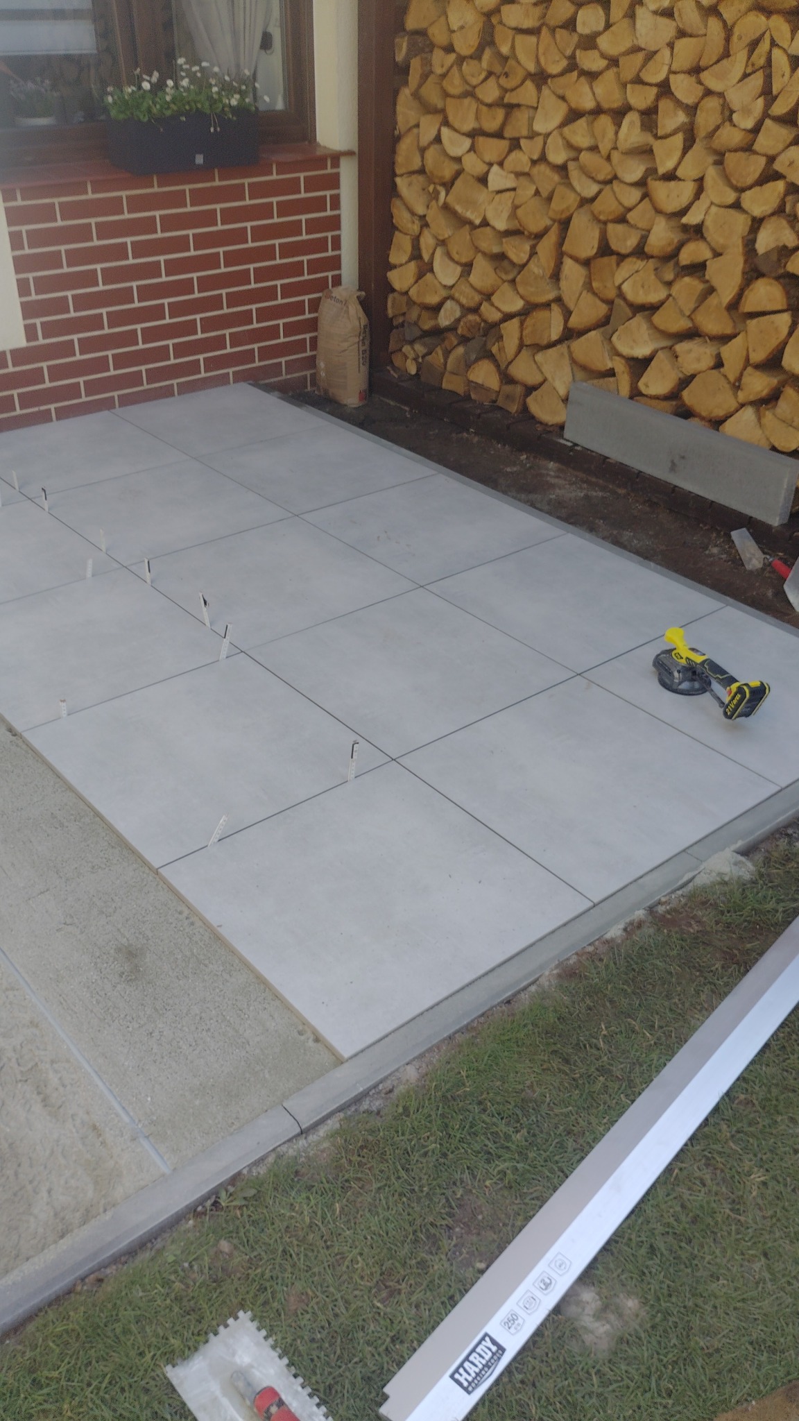 Układanie jasnych płytek na zewnątrz budynku, widoczne narzędzia i krzyżyki dystansowe, przygotowane drewno na opał w tle, częściowo widoczna ściana budynku.