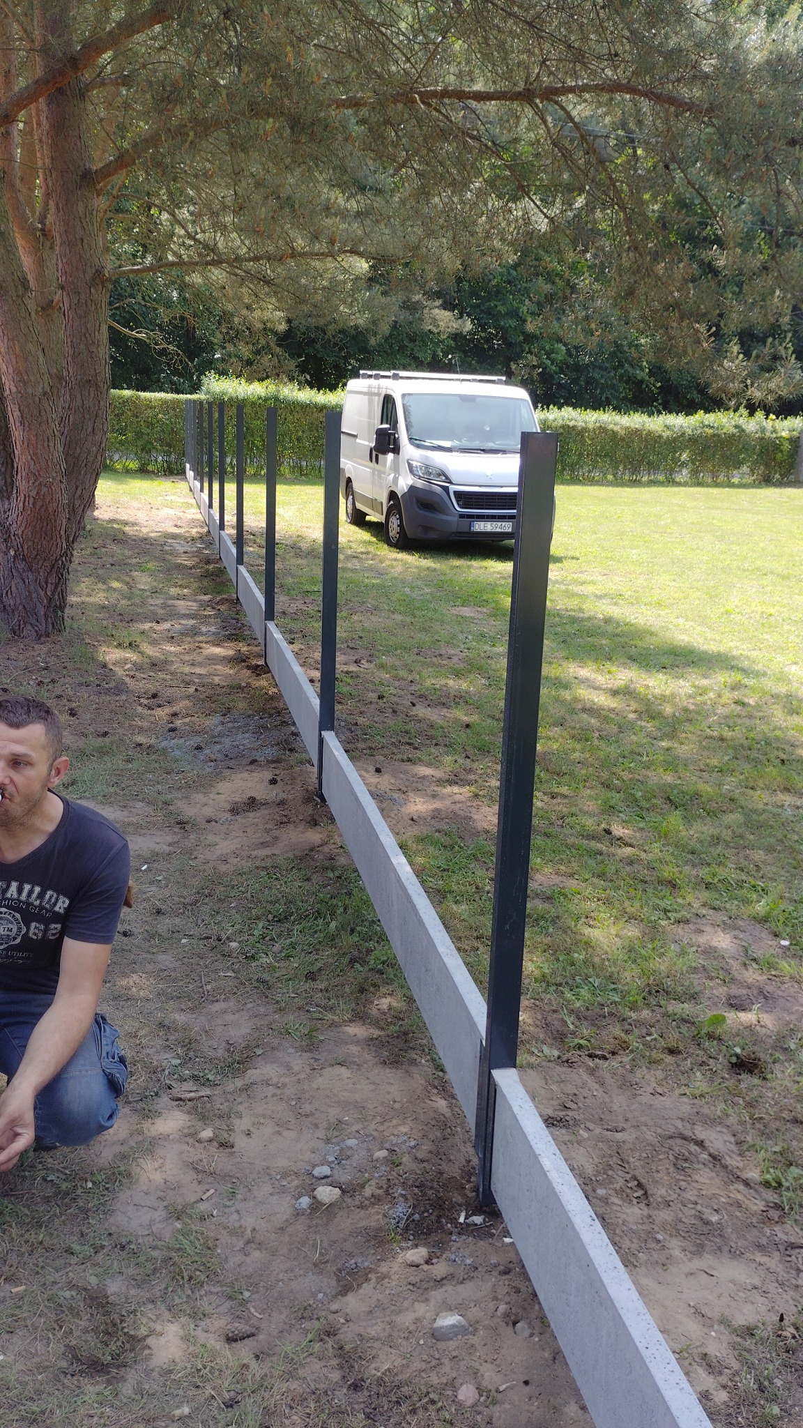 Montaż prefabrykowanego ogrodzenia betonowego z metalowymi słupkami na trawiastym terenie. W tle biały bus i zielony żywopłot.