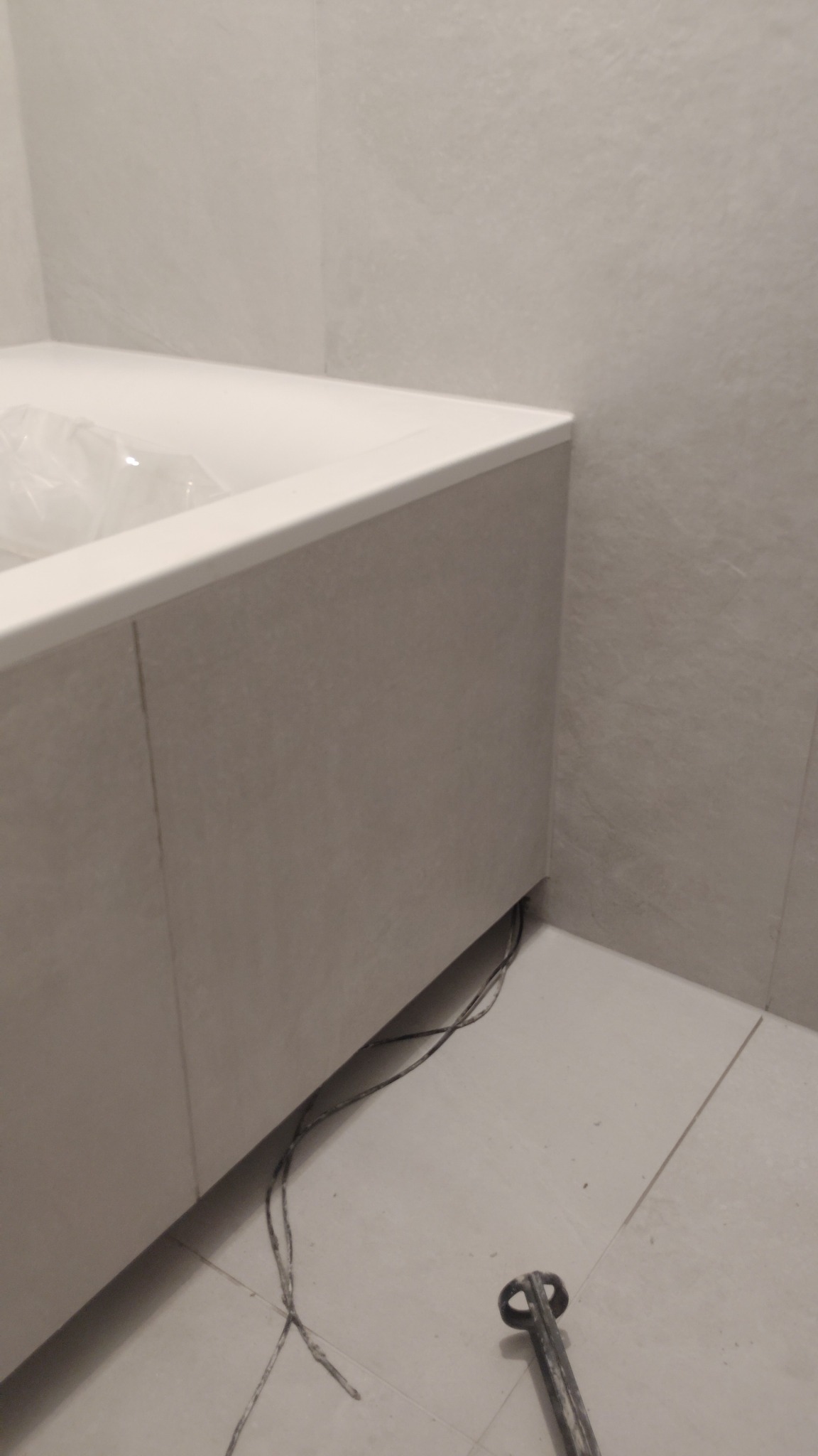 Obudowana wanna z szarą, matową okładziną, widoczne kable i narzędzie na podłodze. Minimalistyczne wykończenie łazienki z akcentem na detale.