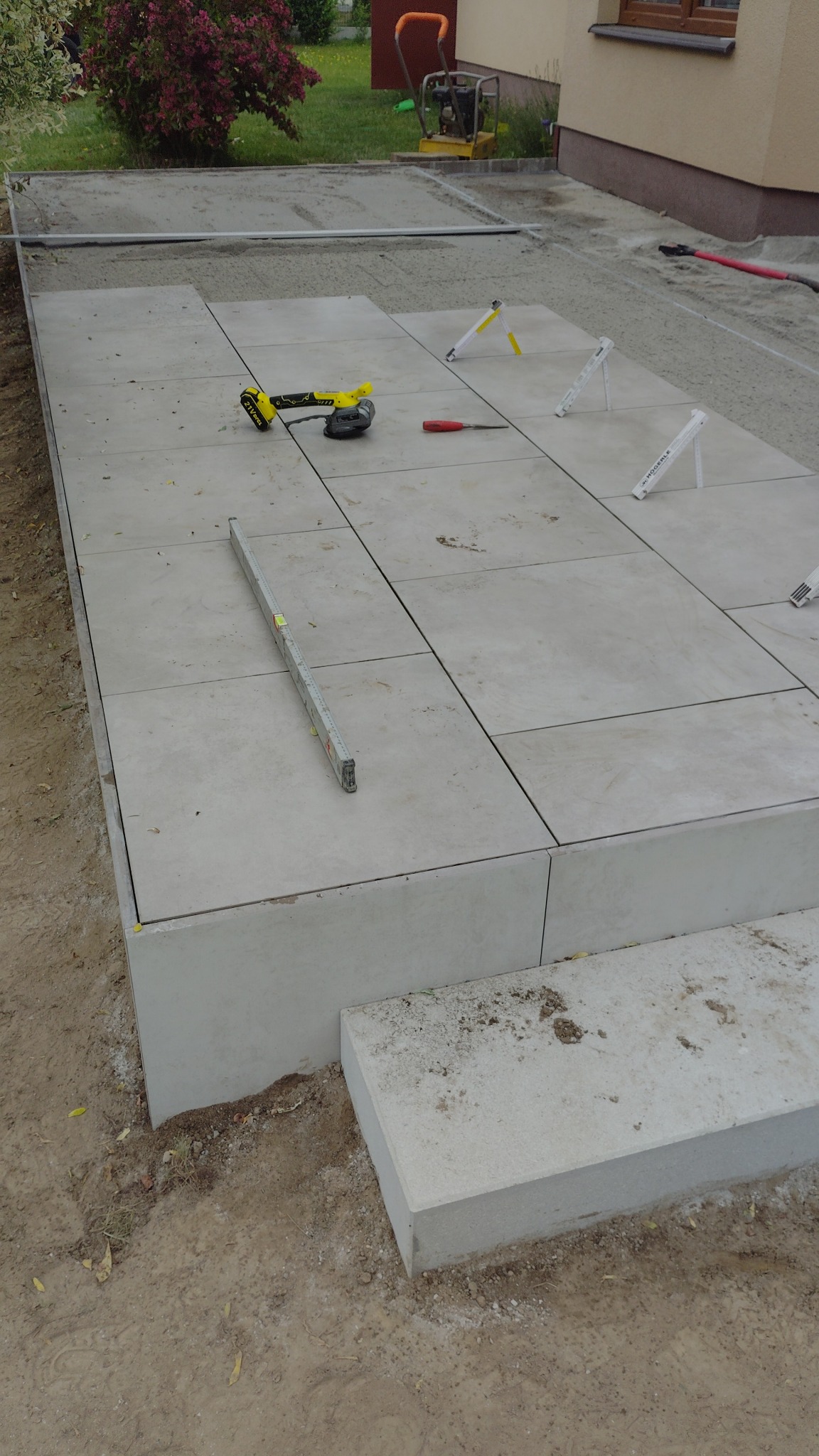 Układanie jasnoszarych płyt chodnikowych na podwyższonym tarasie. Widoczne narzędzia: poziomica, szlifierka kątowa, śrubokręt. W tle zagęszczarka i budynek.