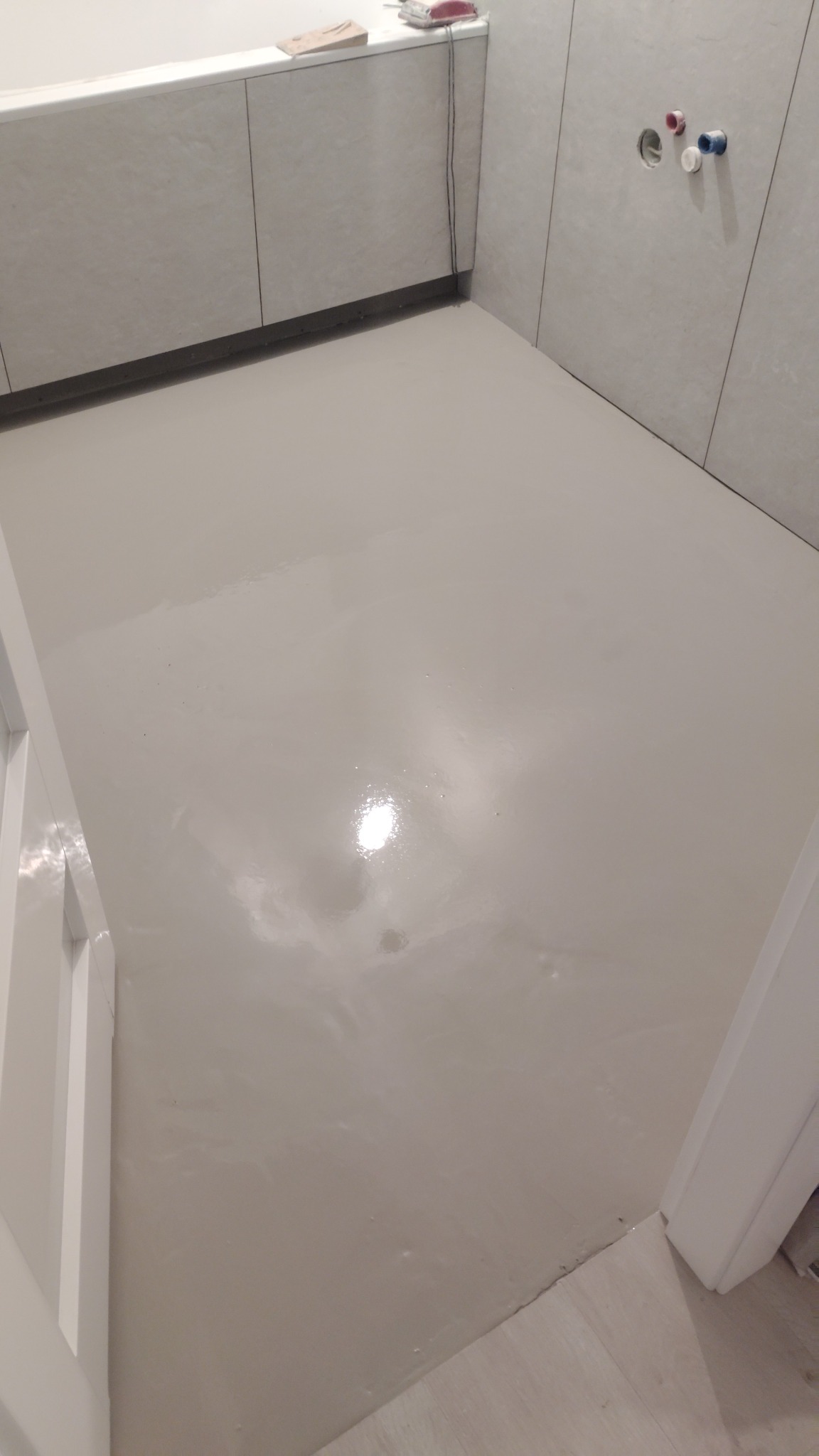 Jasnoszara, gładka wylewka w łazience z białymi płytkami i zabudową wanny. Widoczne przyłącza wodne na ścianie. Minimalistyczne wykończenie podłogi.