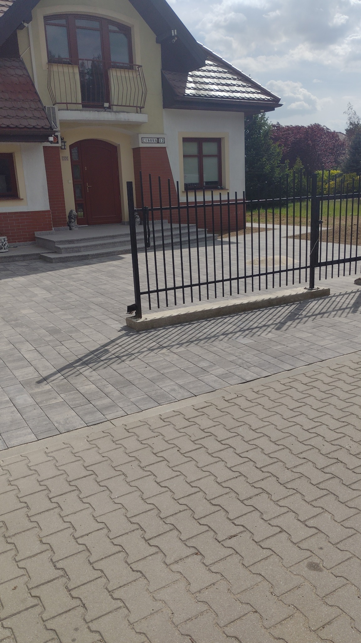 Dwukondygnacyjny dom z balkonem, widoczny zza czarnego ogrodzenia i szarej kostki brukowej. Tabliczka z numerem 'Cisowa 12' nad wejściem.
