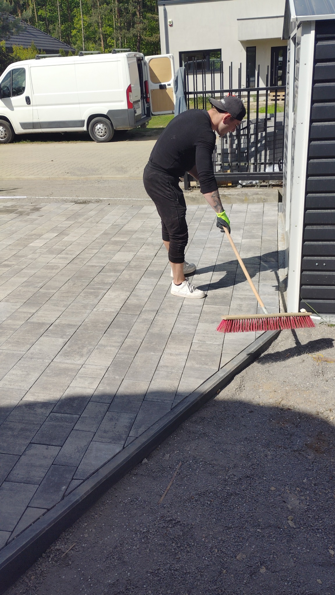Układanie kostki brukowej w Prochowicach: mężczyzna zamiata nową nawierzchnię z szarej kostki, widoczny fragment domu i samochód dostawczy.