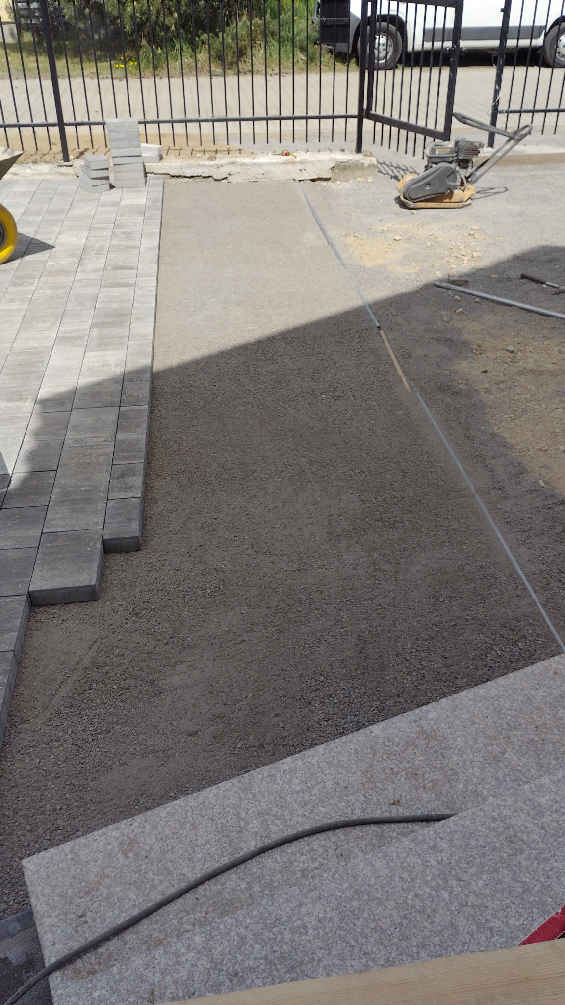 Układanie szarej kostki brukowej na podsypce piaskowej, widoczna ubijarka w tle, ogrodzenie z metalowych prętów.