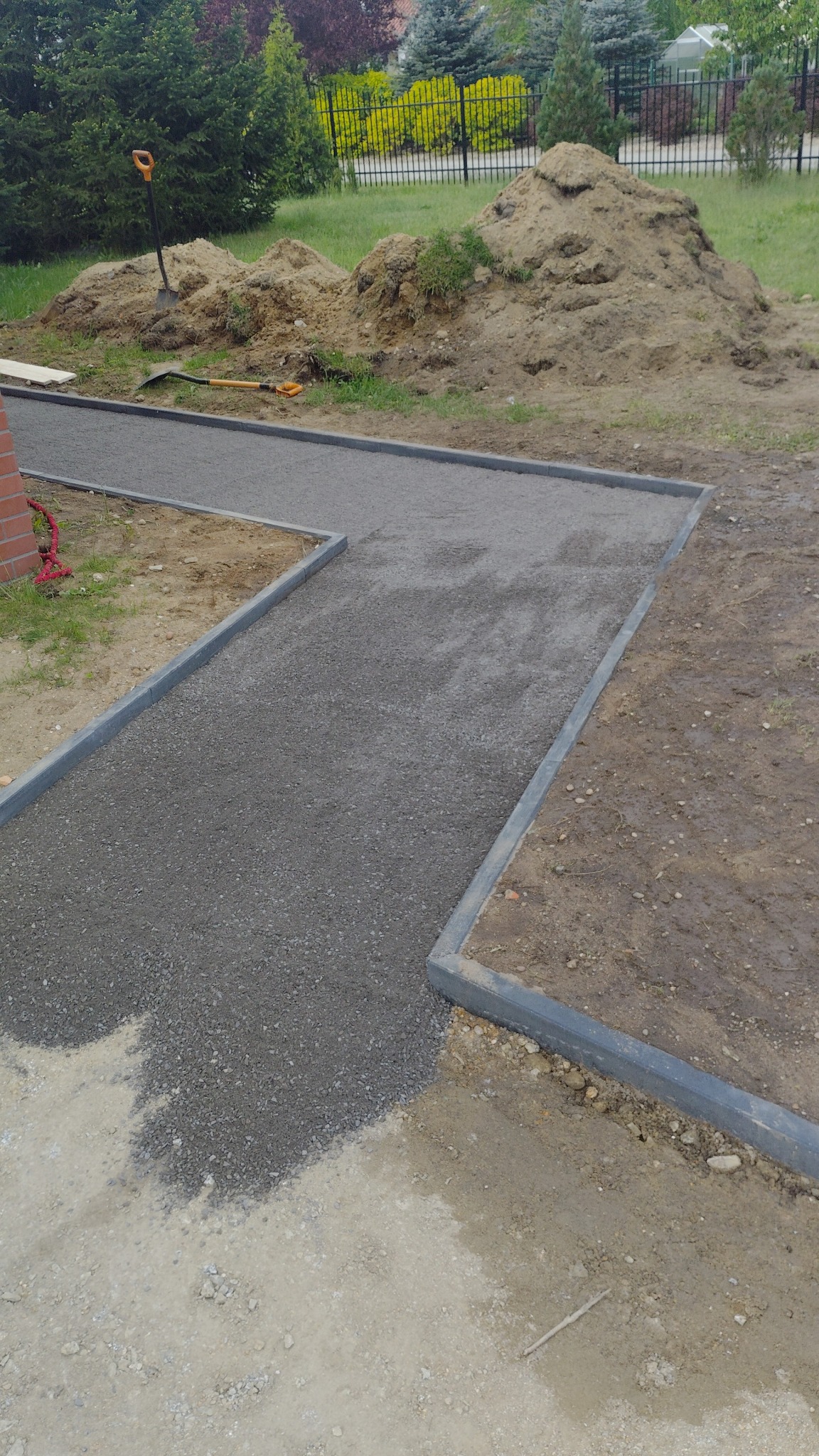 Ścieżka z szarego kruszywa, obramowana betonowymi obrzeżami, w trakcie budowy. W tle hałda ziemi i narzędzia.