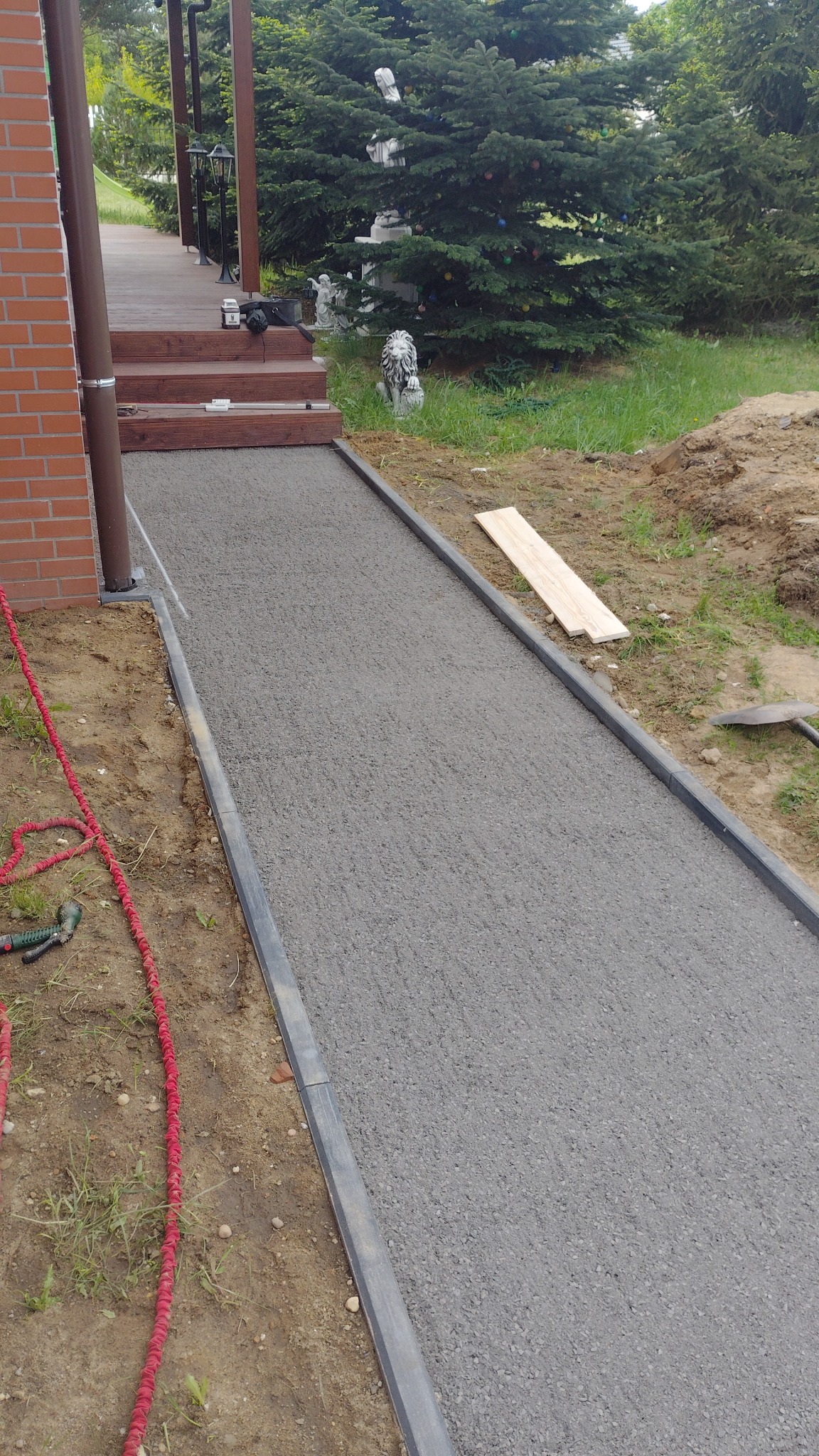 Świeżo ułożona, szara nawierzchnia ścieżki z obrzeżami, prowadząca do drewnianego tarasu z ceglanym budynkiem w tle. Widoczny wąż ogrodowy i narzędzia.