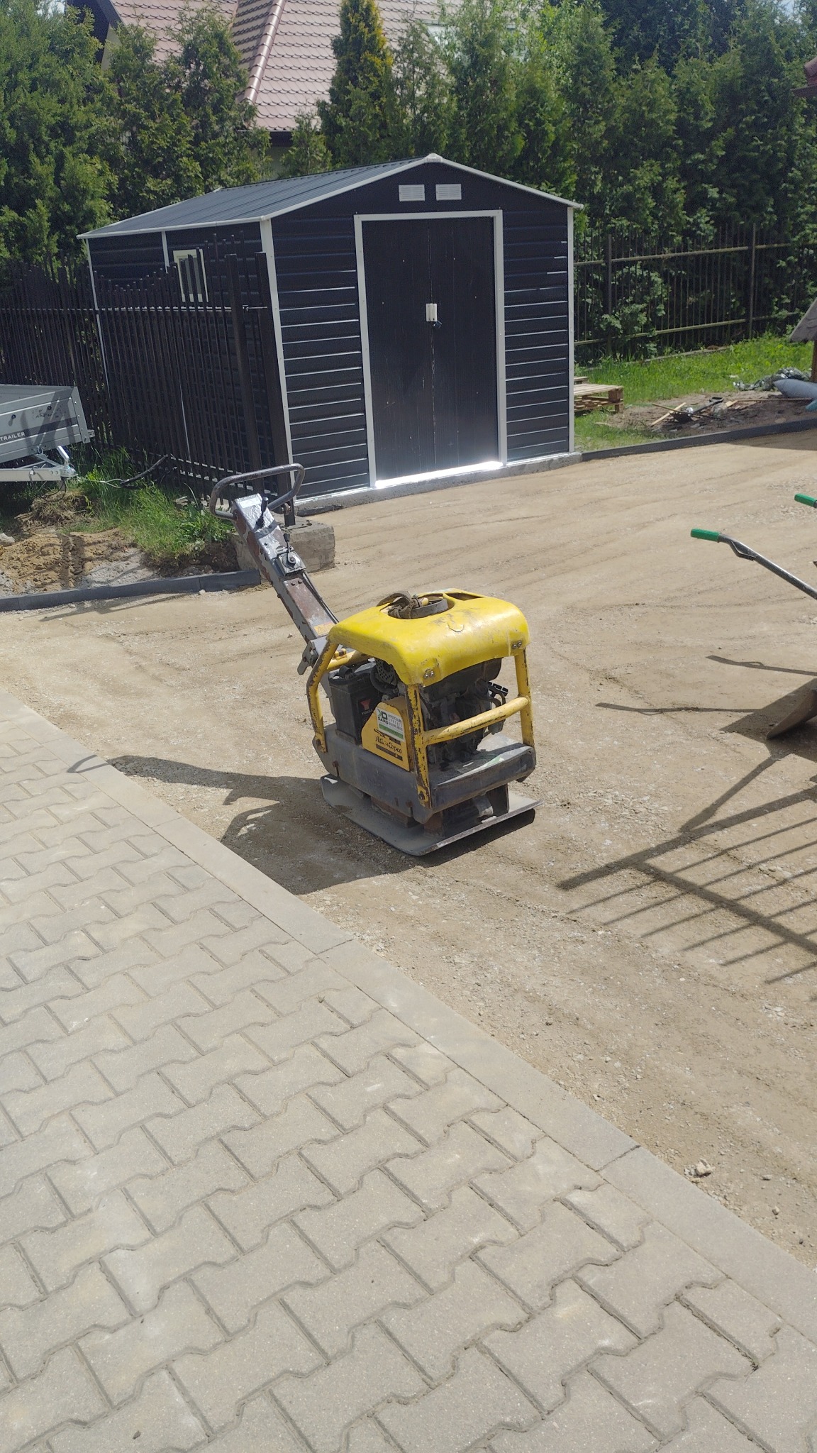 Żółta zagęszczarka na piasku obok kostki brukowej, w tle czarny domek narzędziowy z białymi obramowaniami i ciemne ogrodzenie. Praca w toku, przygotowanie podłoża.