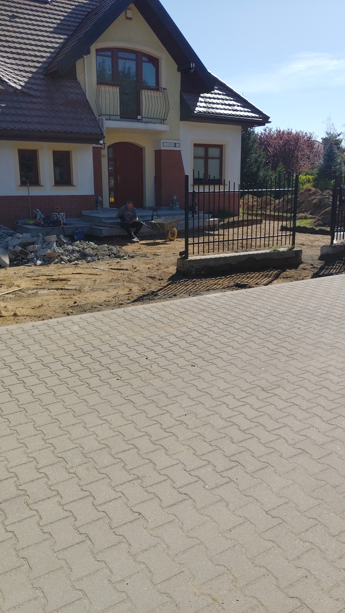 Dom jednorodzinny z numerem 'Cisowa 12' w trakcie prac wykończeniowych na zewnątrz, widoczny pracownik, kostka brukowa i materiały budowlane.