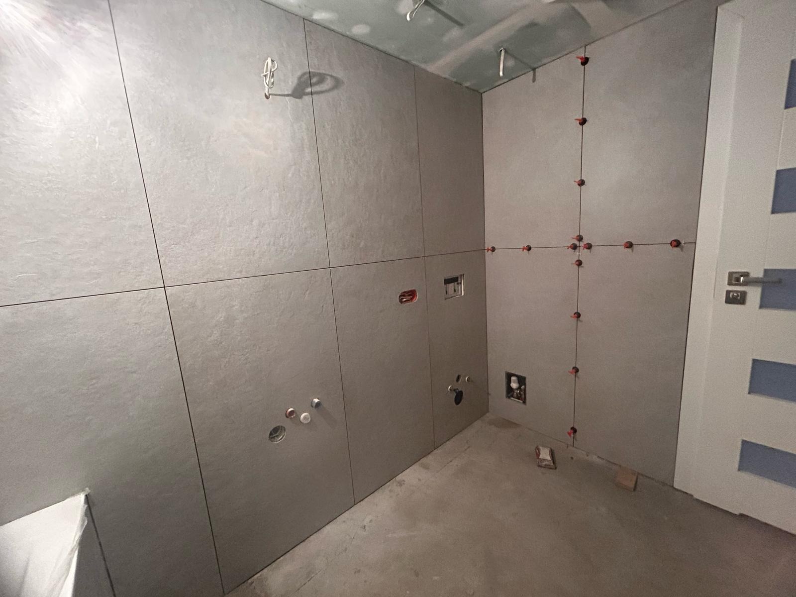 Szare, wielkoformatowe płytki ścienne w trakcie montażu w łazience. Widoczne krzyżyki dystansowe i otwory na instalacje. Surowy beton na podłodze, białe drzwi.