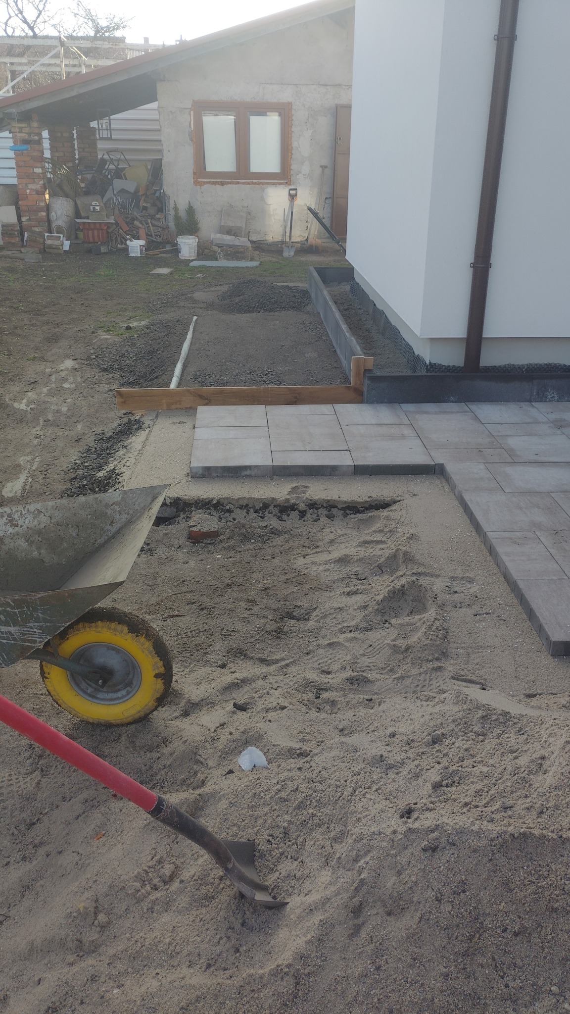 Układanie kostki brukowej na podsypce piaskowej, widoczna taczka i łopata. Częściowo ułożona nawierzchnia przy białej ścianie budynku.