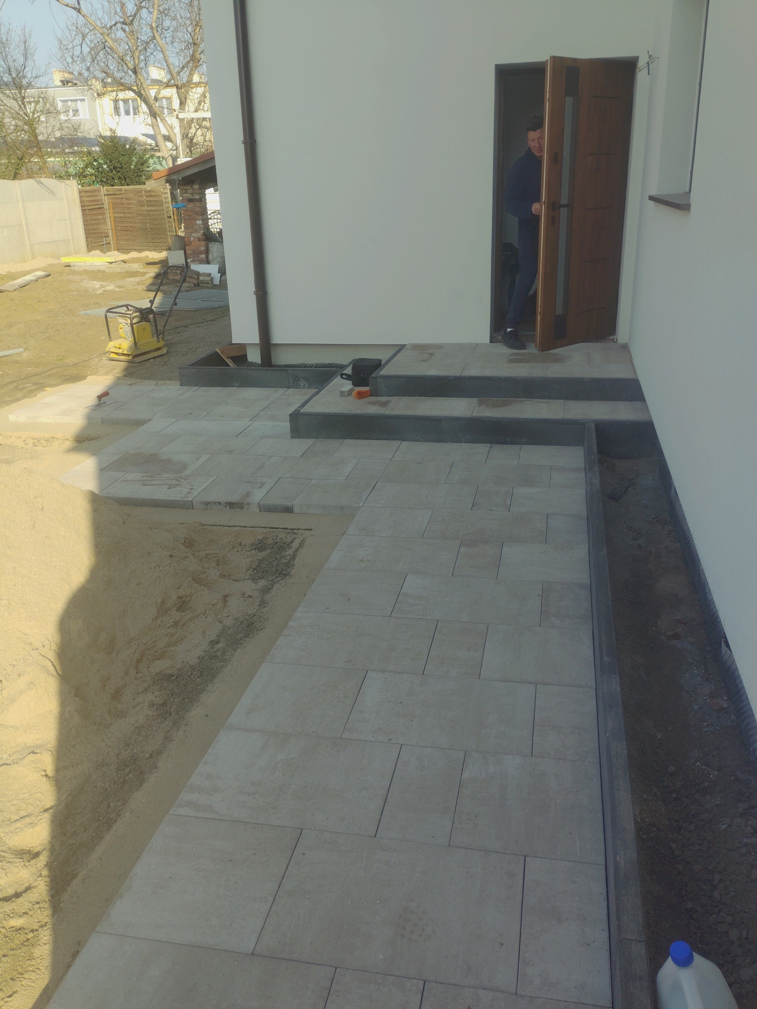 Układanie jasnoszarej kostki brukowej przy białym budynku. Widoczny fragment wejścia z otwartymi drzwiami i osoba w środku. Na ziemi piasek i ubijarka.