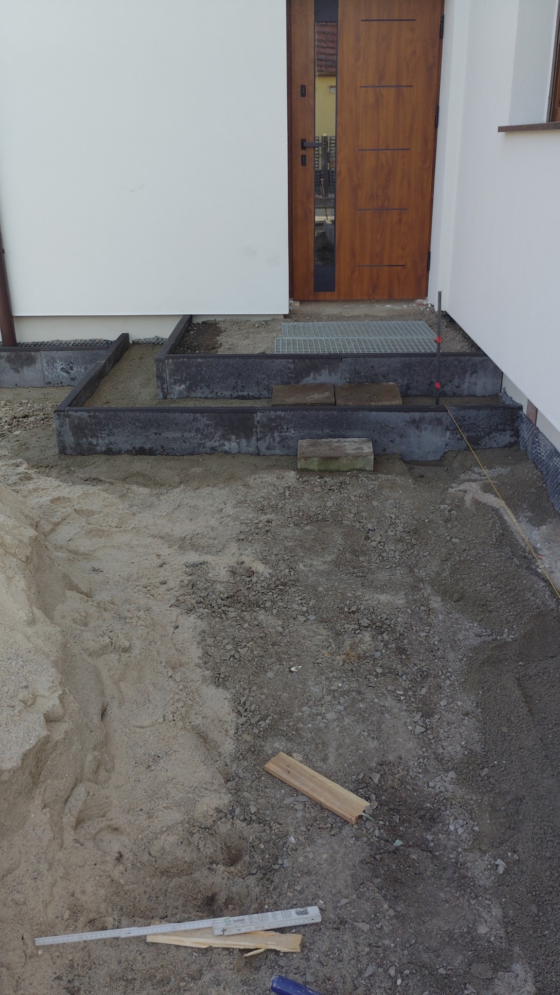 Przygotowanie podłoża pod schody wejściowe do domu. Widoczne obrzeża, piasek, gruz i narzędzia. Etap prac ziemnych przed wykończeniem.