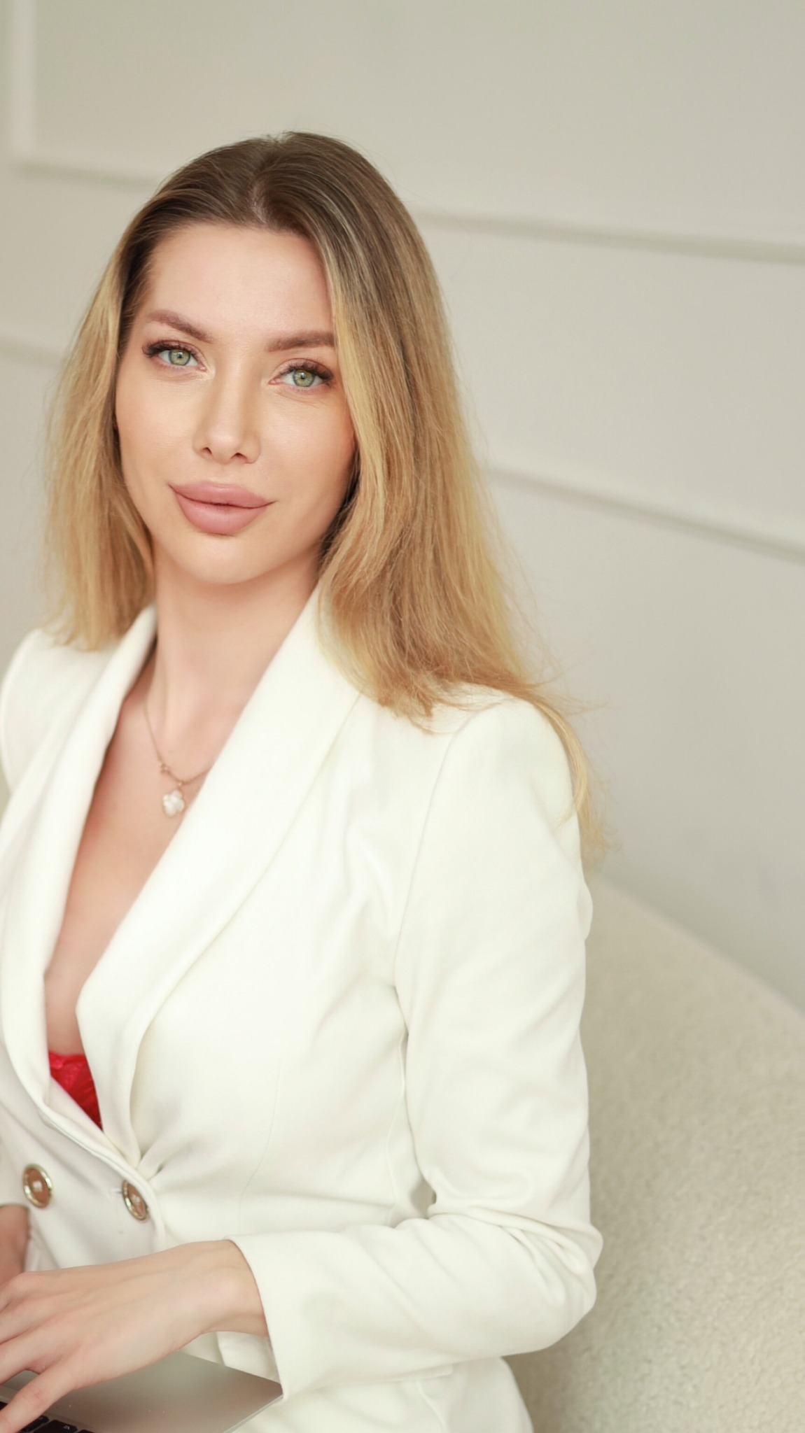 Portret kobiety w białym żakiecie, z delikatnym makijażem i blond włosami, siedzącej na jasnej sofie. Widoczny fragment laptopa.