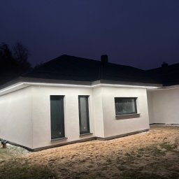 HG-Elektryk SmartTechnika - Nowoczesny dom jednorodzinny z oświetleniem LED pod okapem, sfotografowany nocą. Widoczne okna i elewacja w jasnym kolorze. Na ziemi piasek.