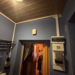 HG-Elektryk SmartTechnika - Szare wnętrze z licznikiem elektrycznym na ścianie, widoczne wejście do innego pomieszczenia z zasłoną z koralików. Oświetlenie sufitowe.