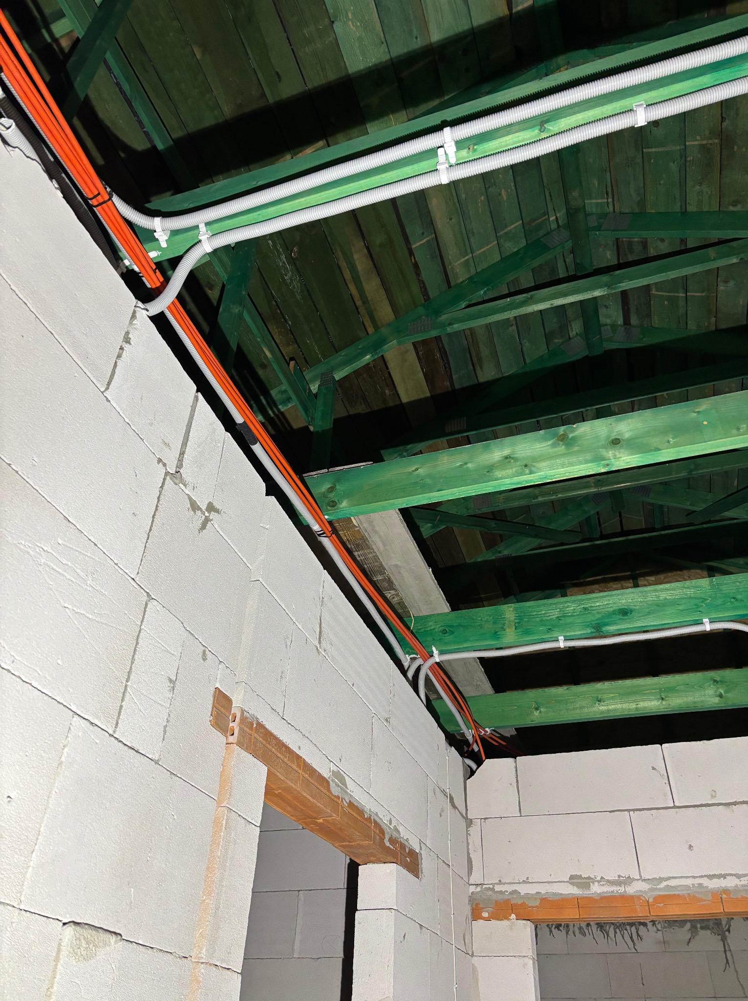 Wnętrze w trakcie budowy z widocznymi, pomarańczowymi i szarymi przewodami elektrycznymi biegnącymi po ścianach z białego betonu komórkowego i zielonym drewnianym stropie.