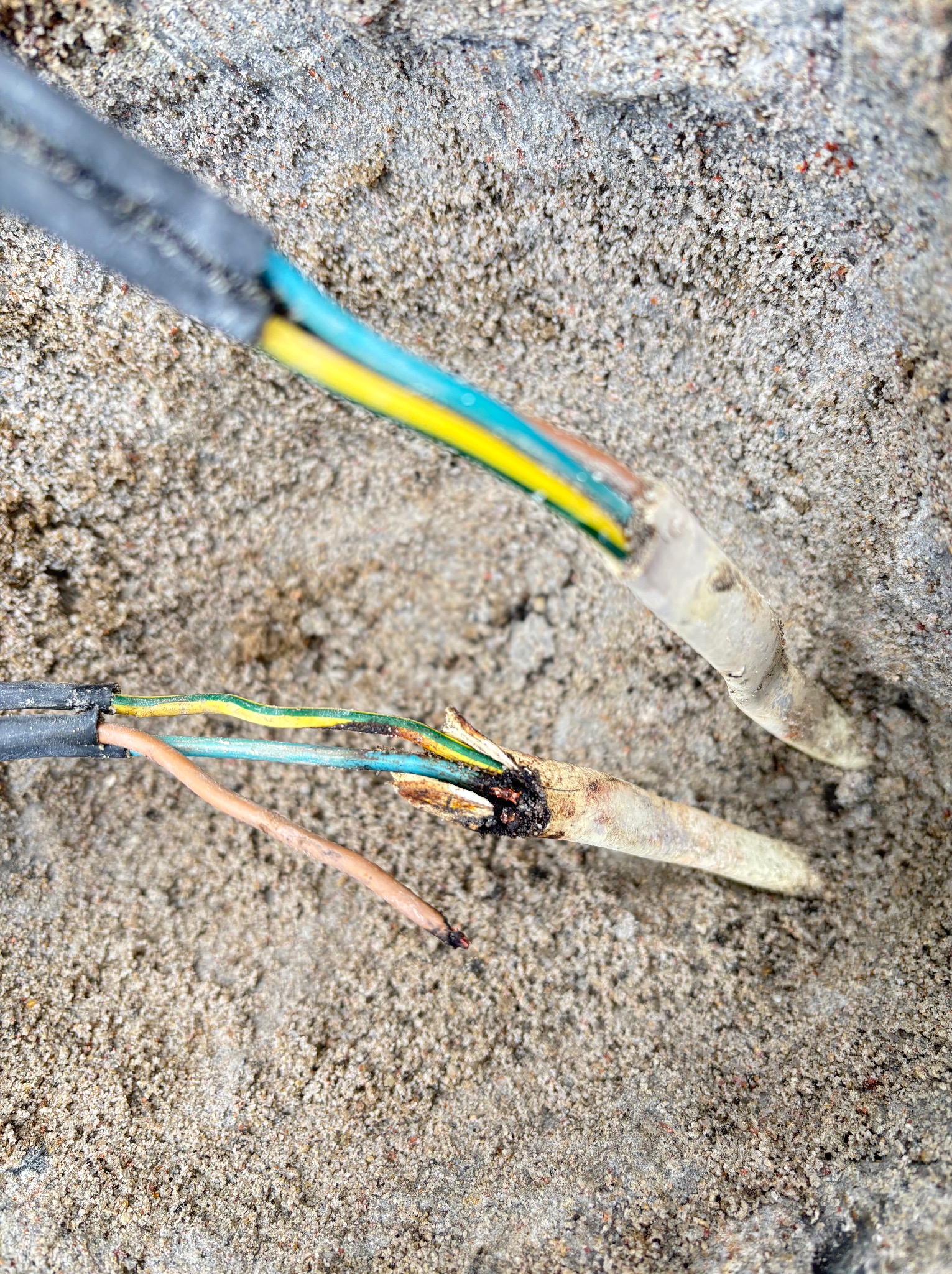 Uszkodzone przewody elektryczne z odsłoniętymi żyłami w żółto-niebiesko-zielonej izolacji, leżące na szorstkiej, betonowej powierzchni. Widoczne ślady przepalenia.