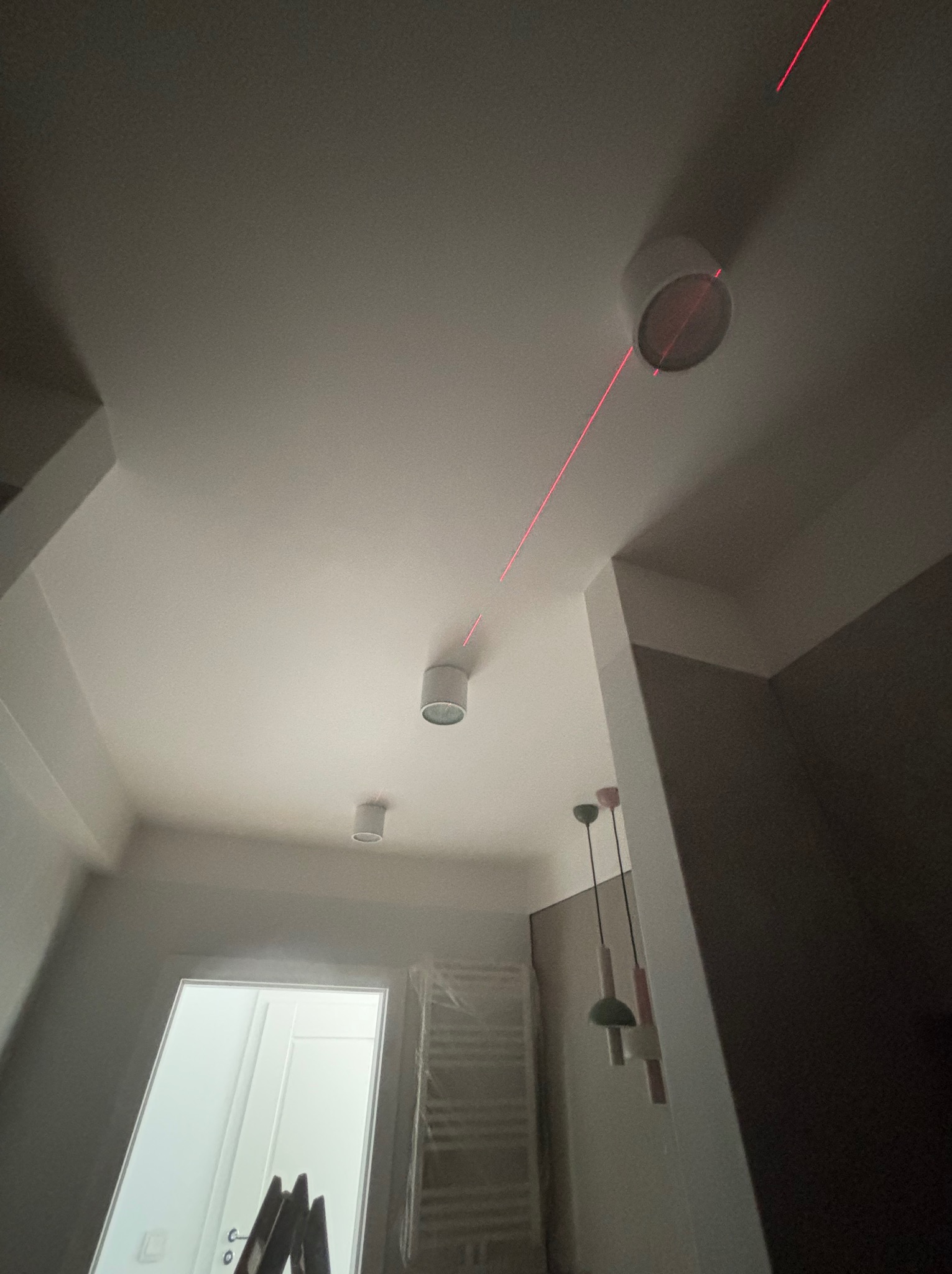 Białe, okrągłe lampy sufitowe zamontowane w jasnym pomieszczeniu, wyznaczane czerwoną linią lasera. Widoczna drabina i fragment grzejnika.