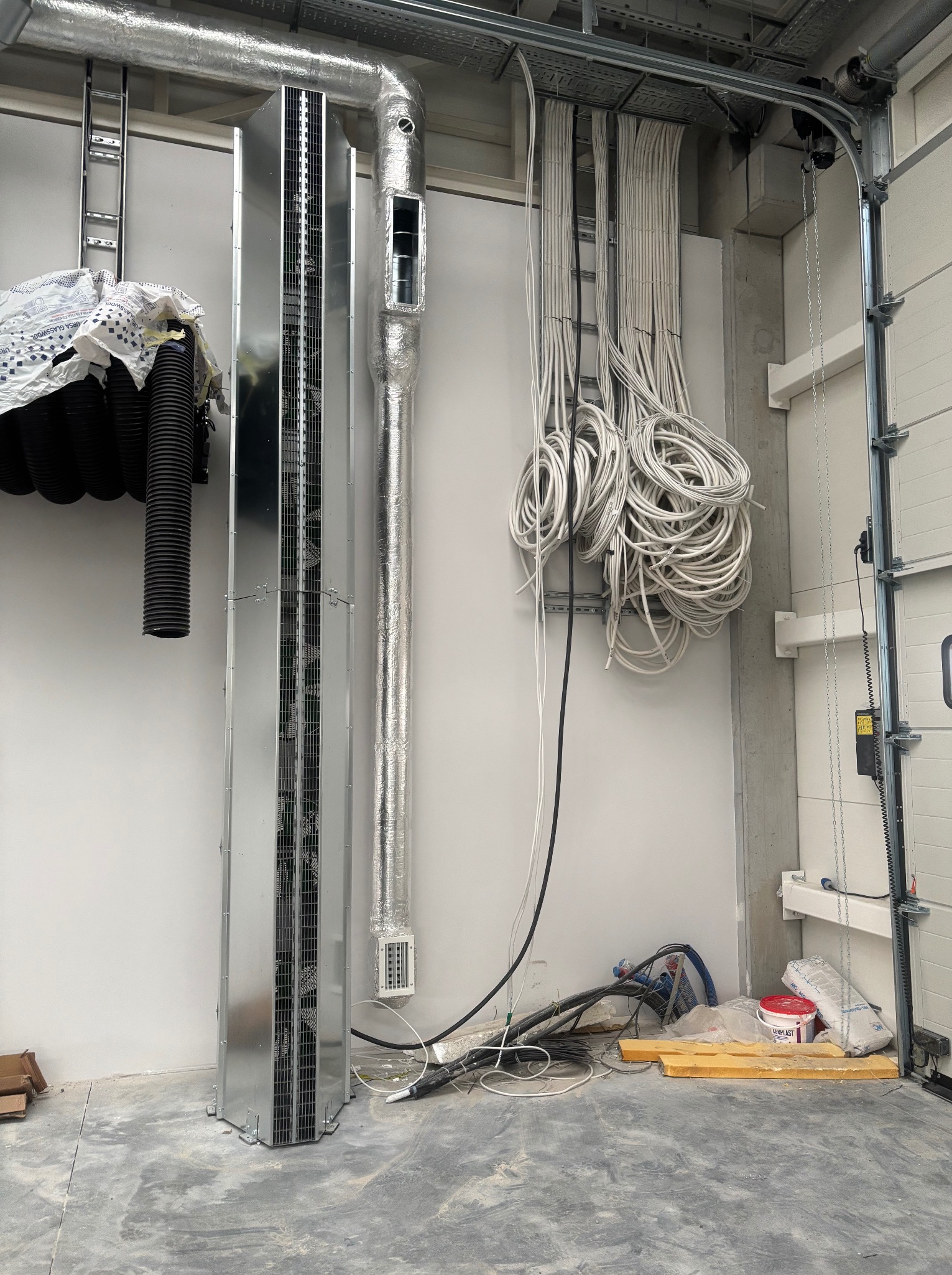 Wnętrze hali z widoczną instalacją wentylacyjną, korytami kablowymi i zwisającymi przewodami elektrycznymi. Widoczne elementy montażowe i materiały budowlane.