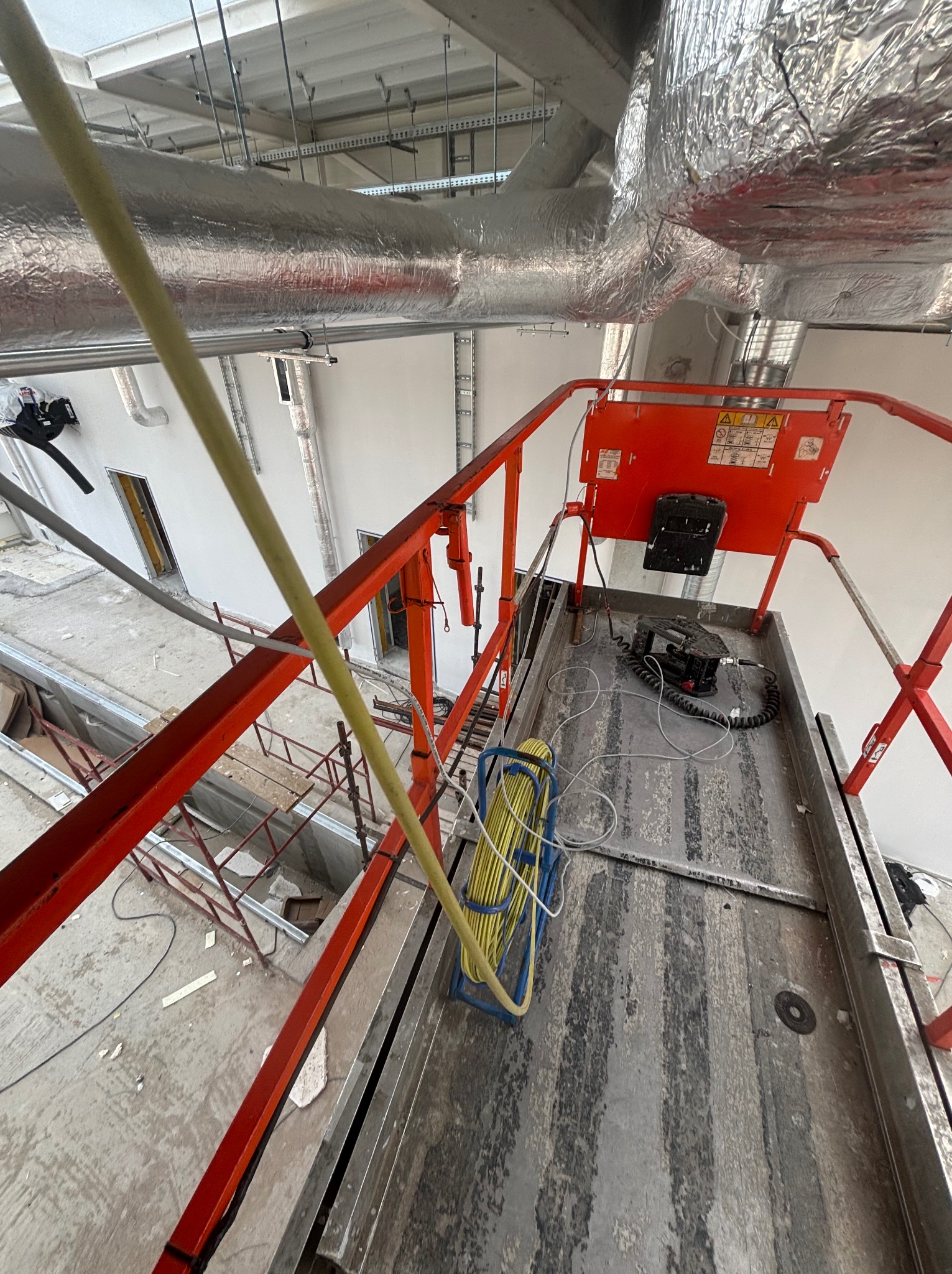 Wysięgnik z narzędziami elektrycznymi na budowie, widok z góry. Żółty kabel zwinięty w niebieskim stojaku. Instalacje pod sufitem w tle.