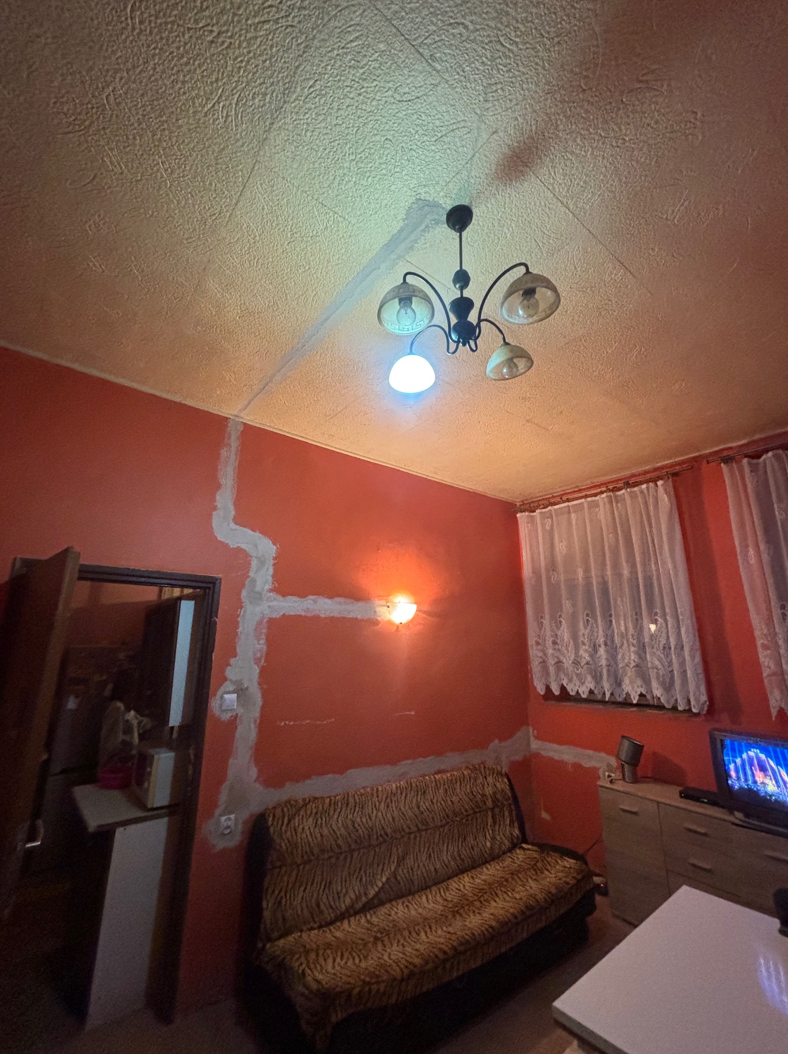 Pokój z pomarańczowymi ścianami, na których widać ślady po bruzdach pod kable, przygotowane do położenia instalacji. Widoczny żyrandol i meble.
