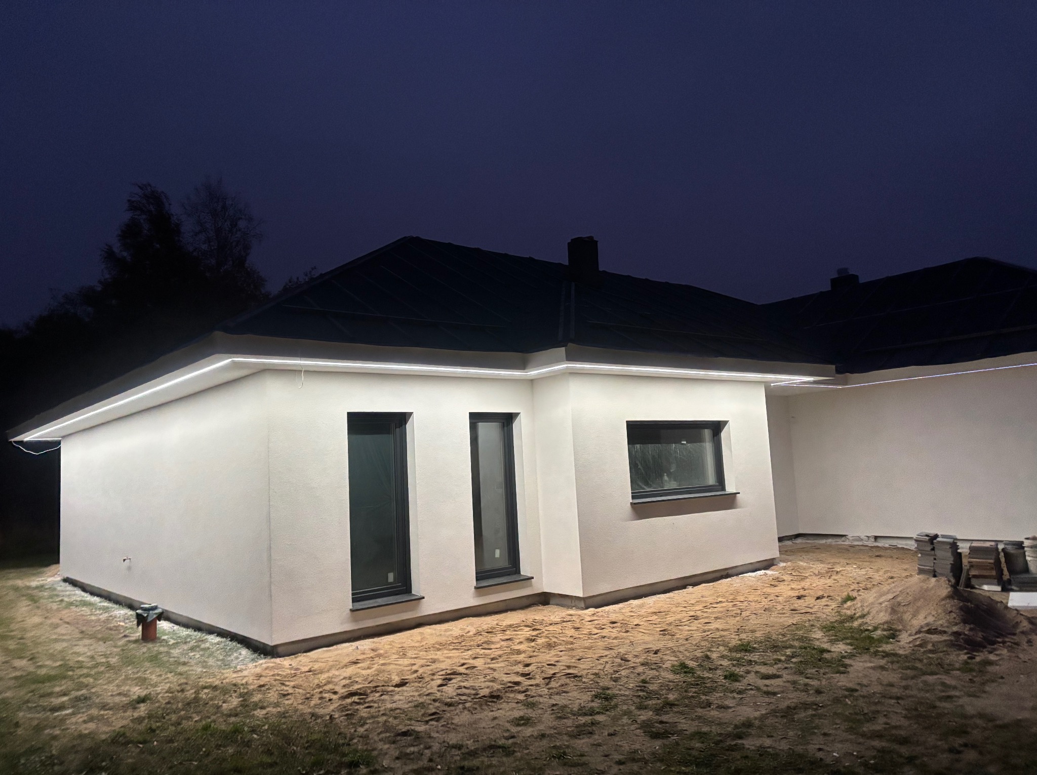 Nowoczesny dom jednorodzinny z oświetleniem LED pod okapem, sfotografowany nocą. Widoczne okna i elewacja w jasnym kolorze. Na ziemi piasek.