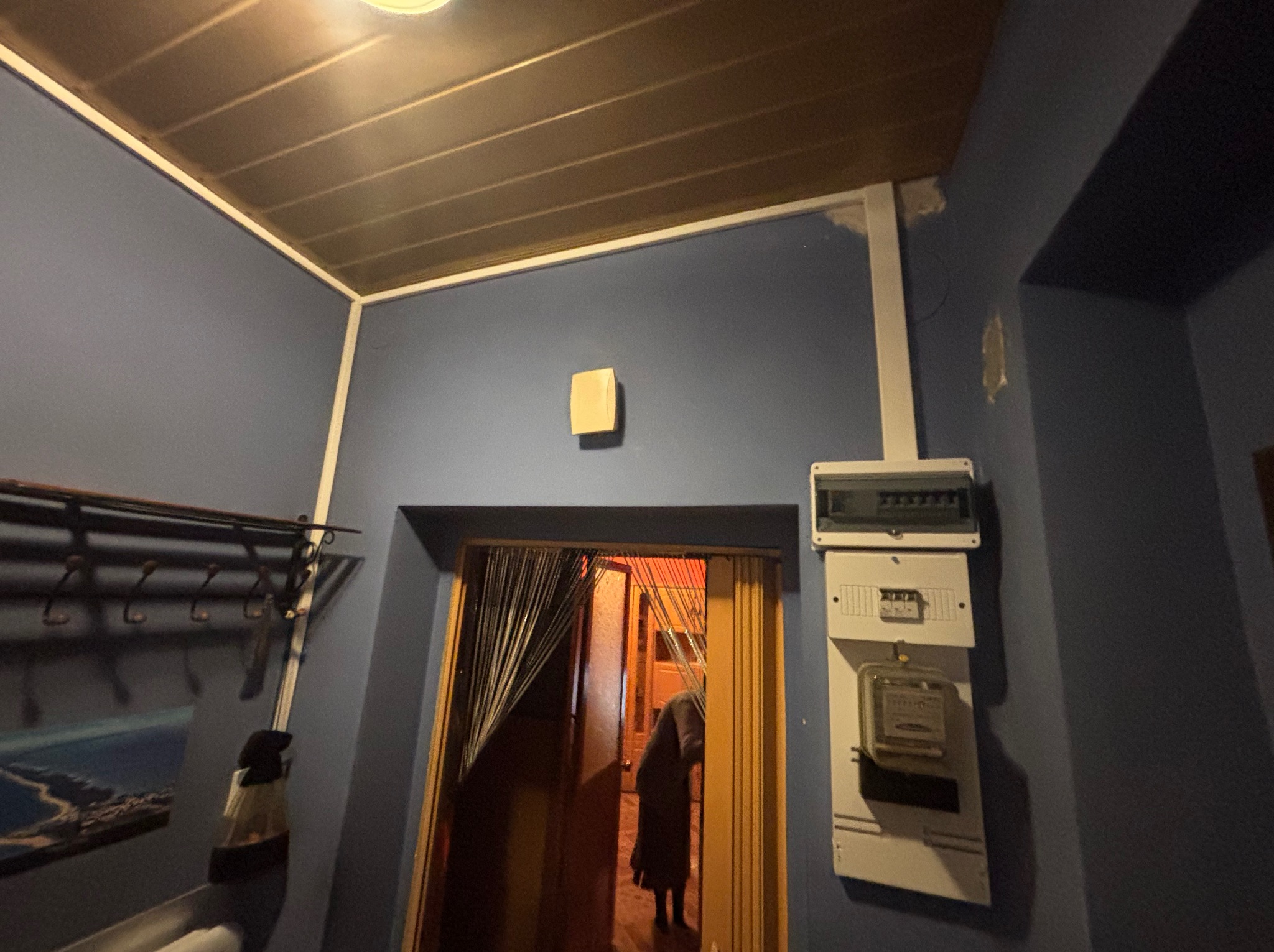 Szare wnętrze z licznikiem elektrycznym na ścianie, widoczne wejście do innego pomieszczenia z zasłoną z koralików. Oświetlenie sufitowe.