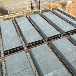 Magbud - Ułożone szare bloczki stropowe Teriva zbrojone stalą, widok z góry na budowie. W tle cegły i brama.