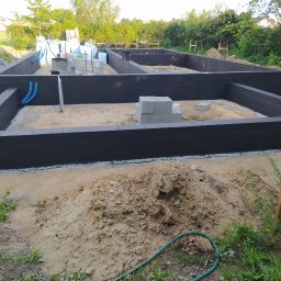 Magbud - Zalane fundamenty domu z wyprowadzonymi rurami instalacji, widoczne bloczki betonowe i materiały izolacyjne. Ujęcie z poziomu gruntu, w tle zieleń i zabudowa.