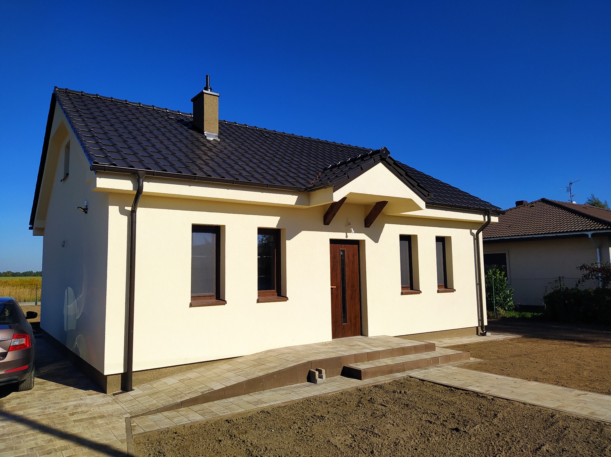 Nowoczesny, parterowy dom z beżową elewacją, ciemną dachówką i brązowymi oknami. Widoczny podjazd dla wózków i świeżo zagospodarowany teren wokół budynku.