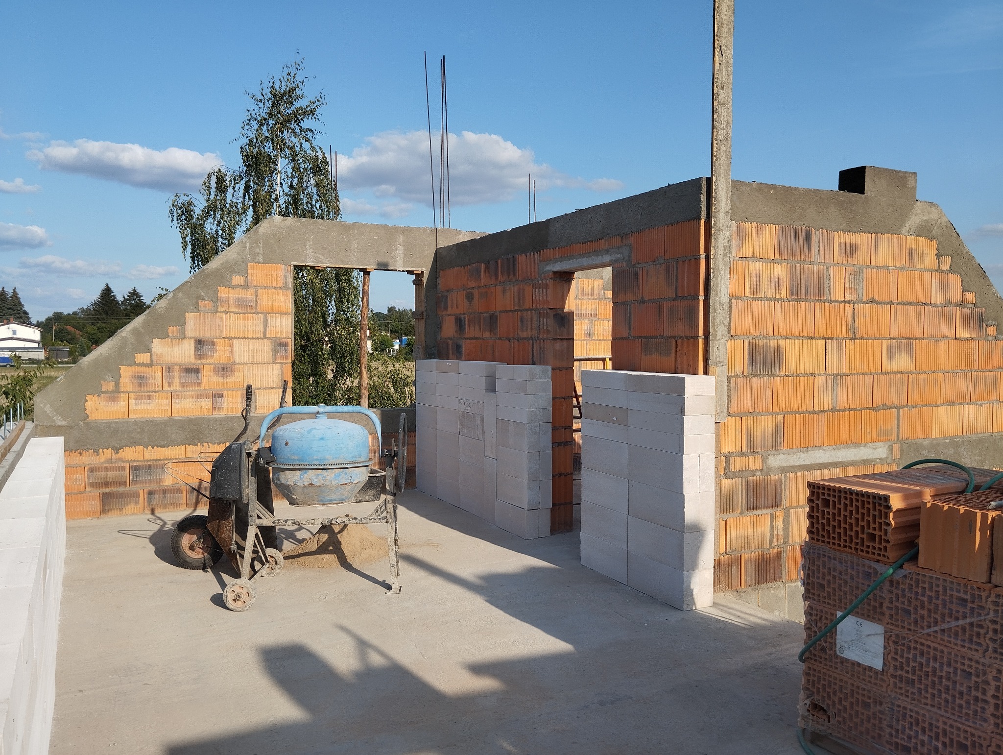 Wznoszenie ścian domu z czerwonej cegły, widoczna betoniarka i białe bloczki. Budowa w toku, zarys okien i drzwi. Słoneczny dzień.