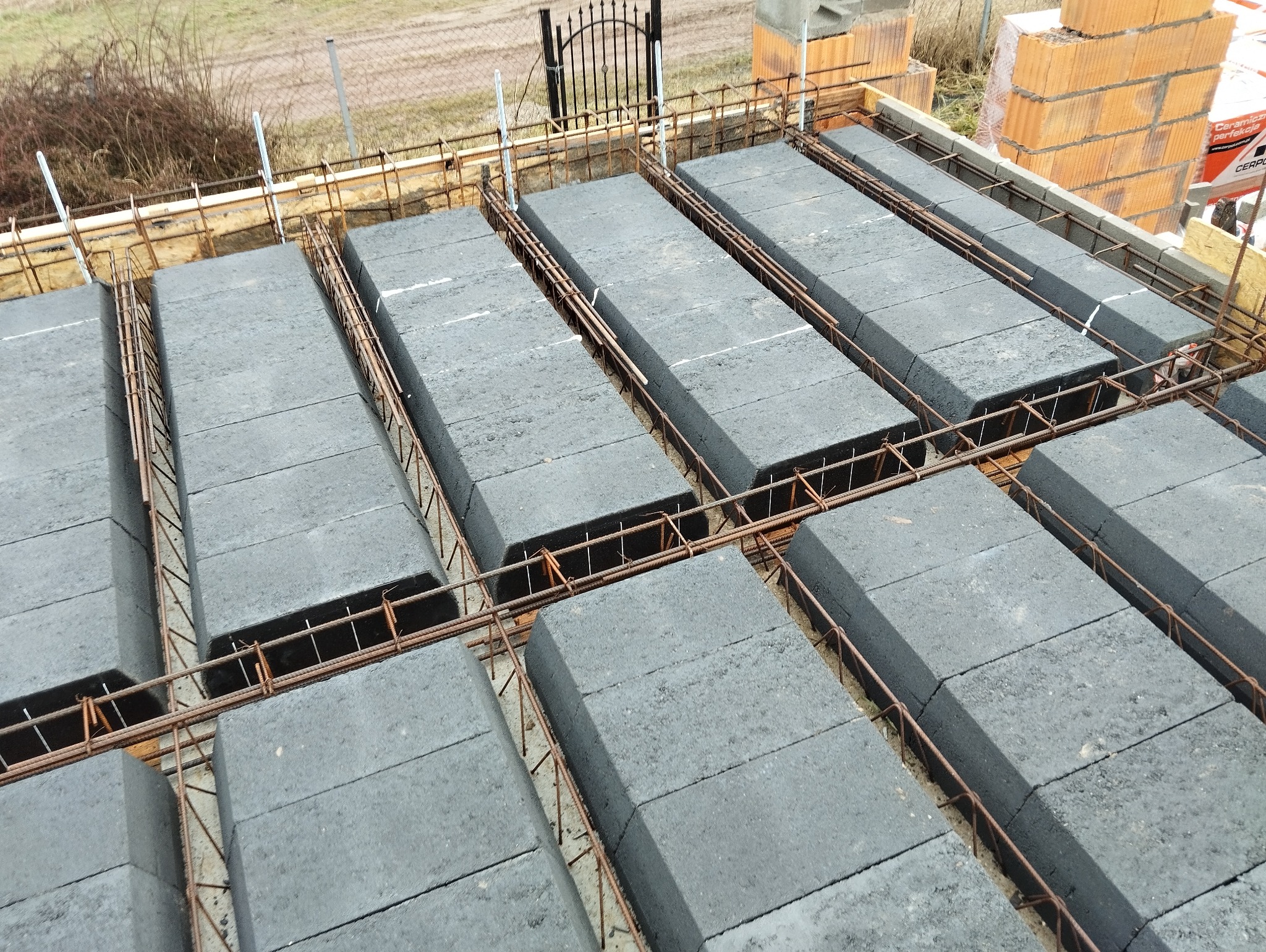 Ułożone szare bloczki stropowe Teriva zbrojone stalą, widok z góry na budowie. W tle cegły i brama.