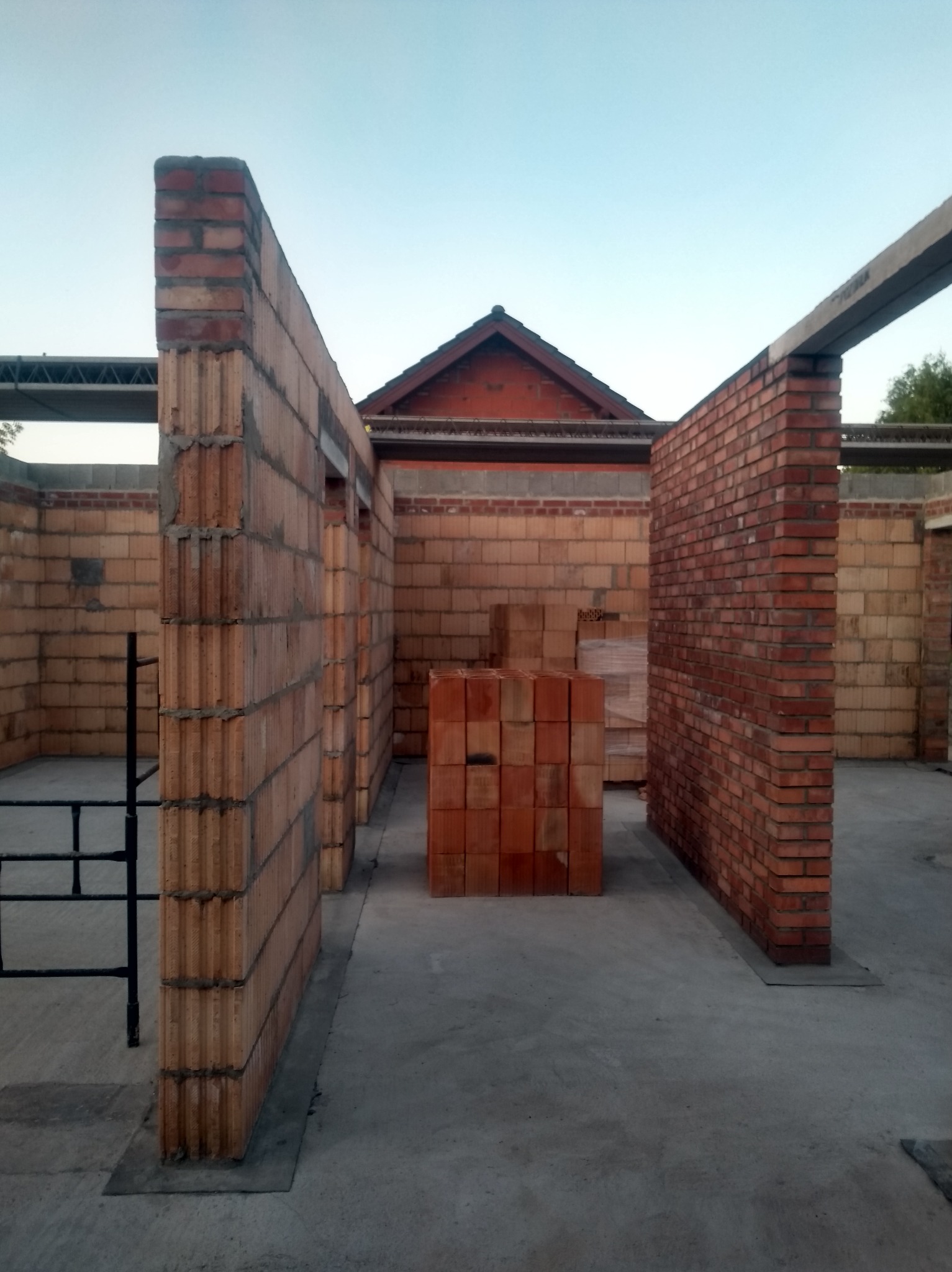 Wnętrze w trakcie budowy z ceglanymi ścianami i blokami, widok na dach budynku w tle. Surowy stan, betonowa podłoga, elementy konstrukcyjne.