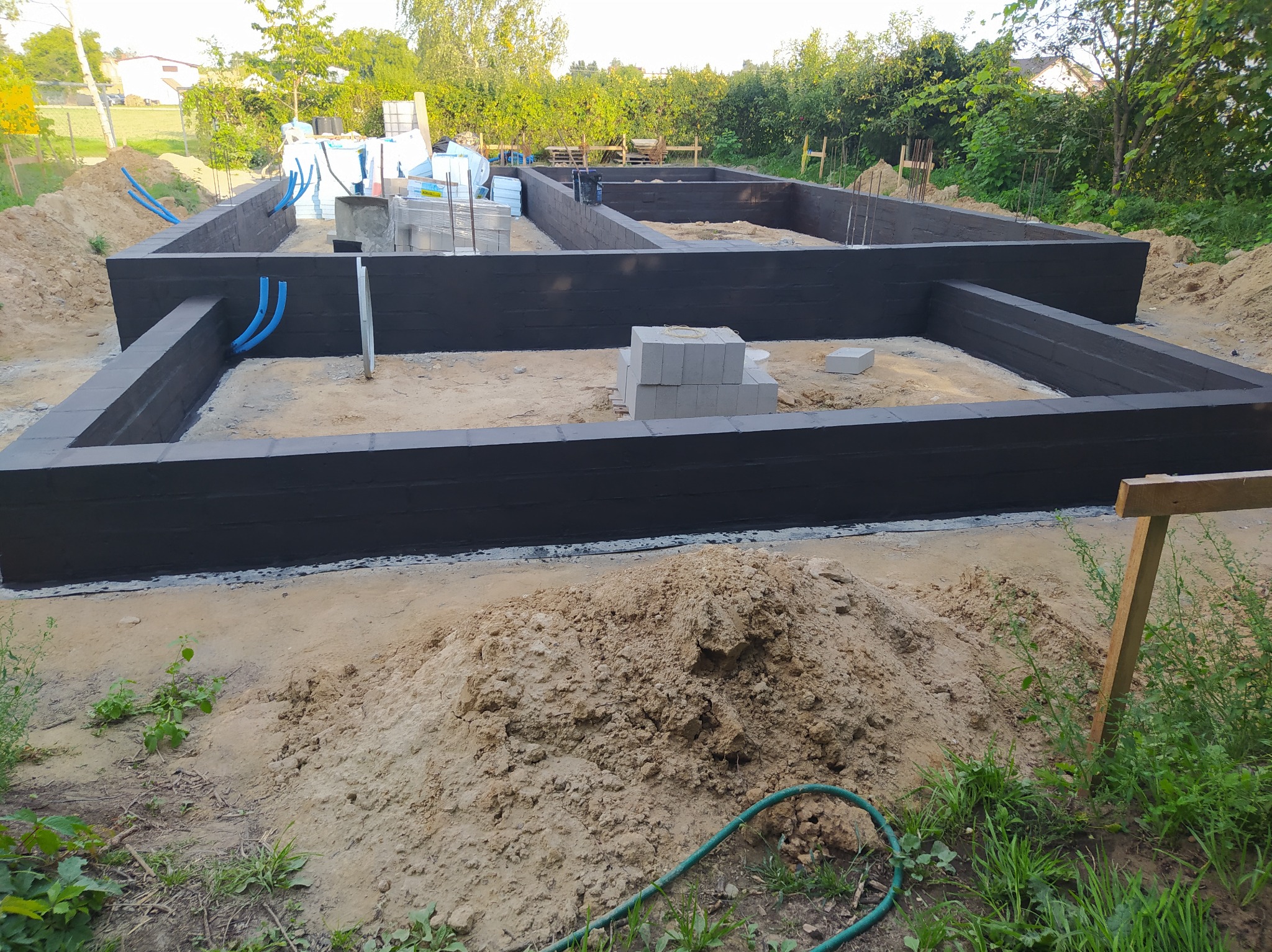 Zalane fundamenty domu z wyprowadzonymi rurami instalacji, widoczne bloczki betonowe i materiały izolacyjne. Ujęcie z poziomu gruntu, w tle zieleń i zabudowa.