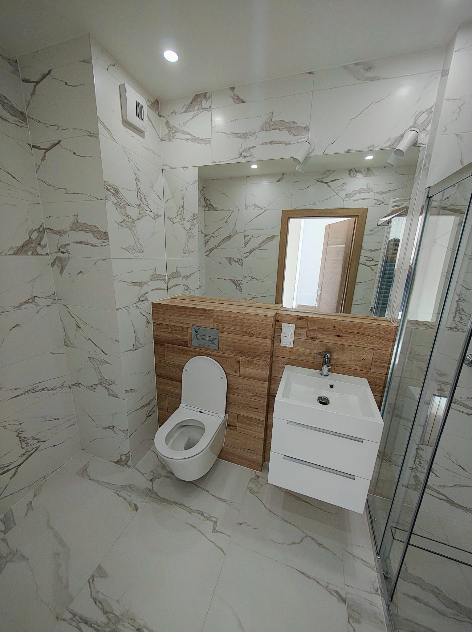 Nowoczesna łazienka z marmurowymi płytkami, drewnianą zabudową WC i umywalką z szafką. Lustro odbija fragment innego pomieszczenia. Minimalistyczny design.