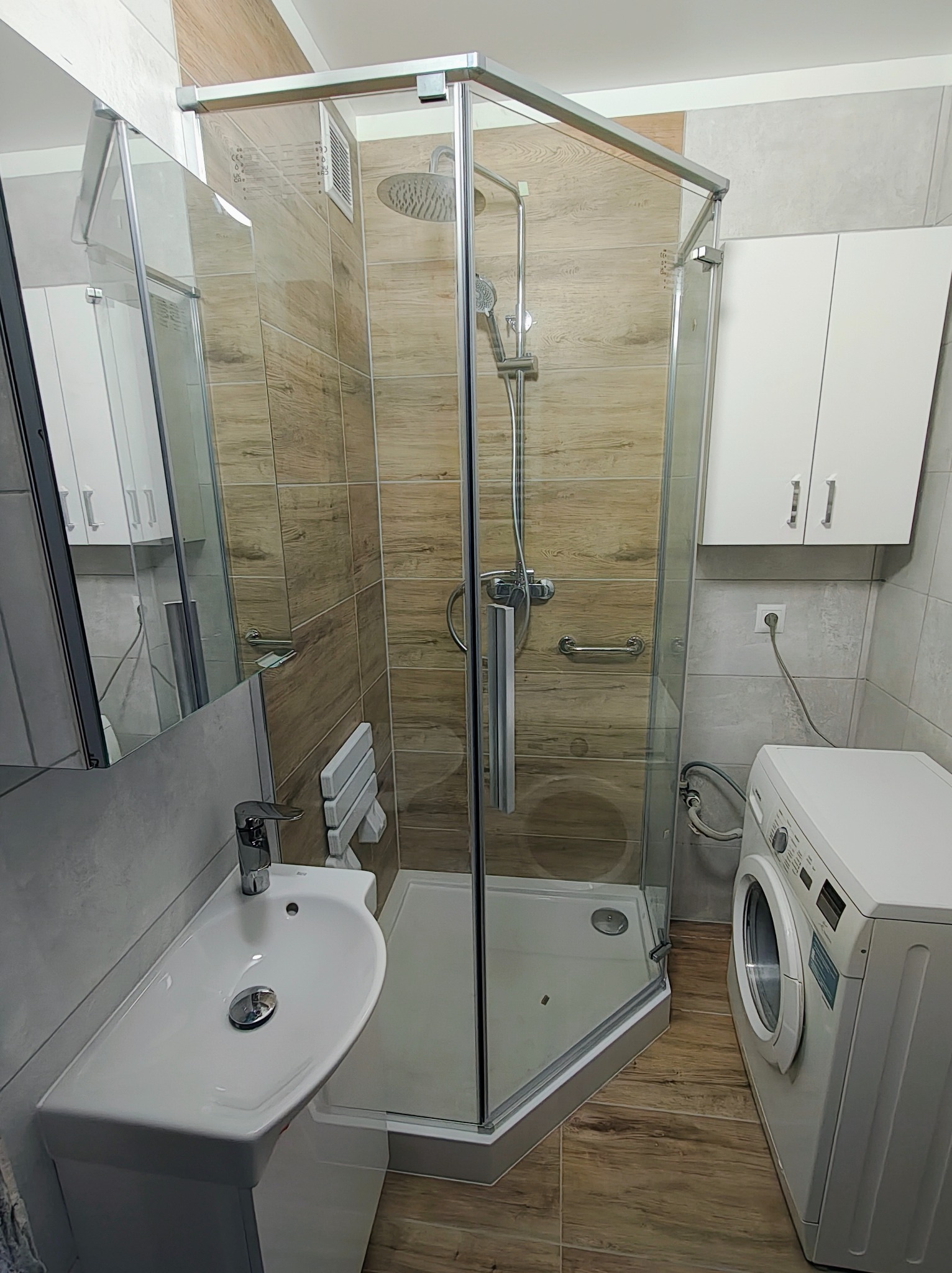 Nowoczesna łazienka po remoncie: narożna kabina prysznicowa z deszczownicą, umywalka z szafką, lustro i pralka. Jasne płytki imitujące drewno na ścianach.
