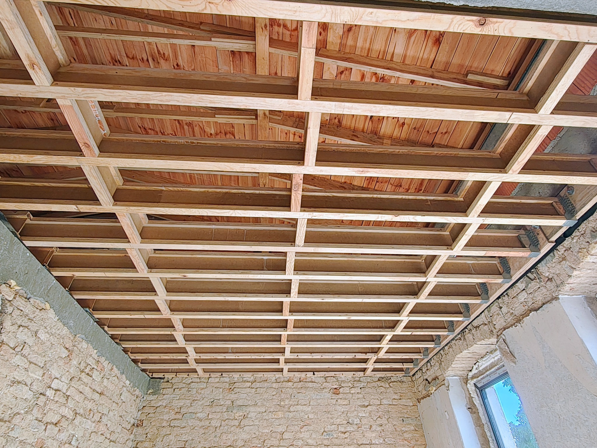 Drewniana konstrukcja sufitu podwieszanego w trakcie montażu, widoczna na tle ceglanej ściany. Elementy konstrukcyjne z jasnego drewna tworzą regularną siatkę.