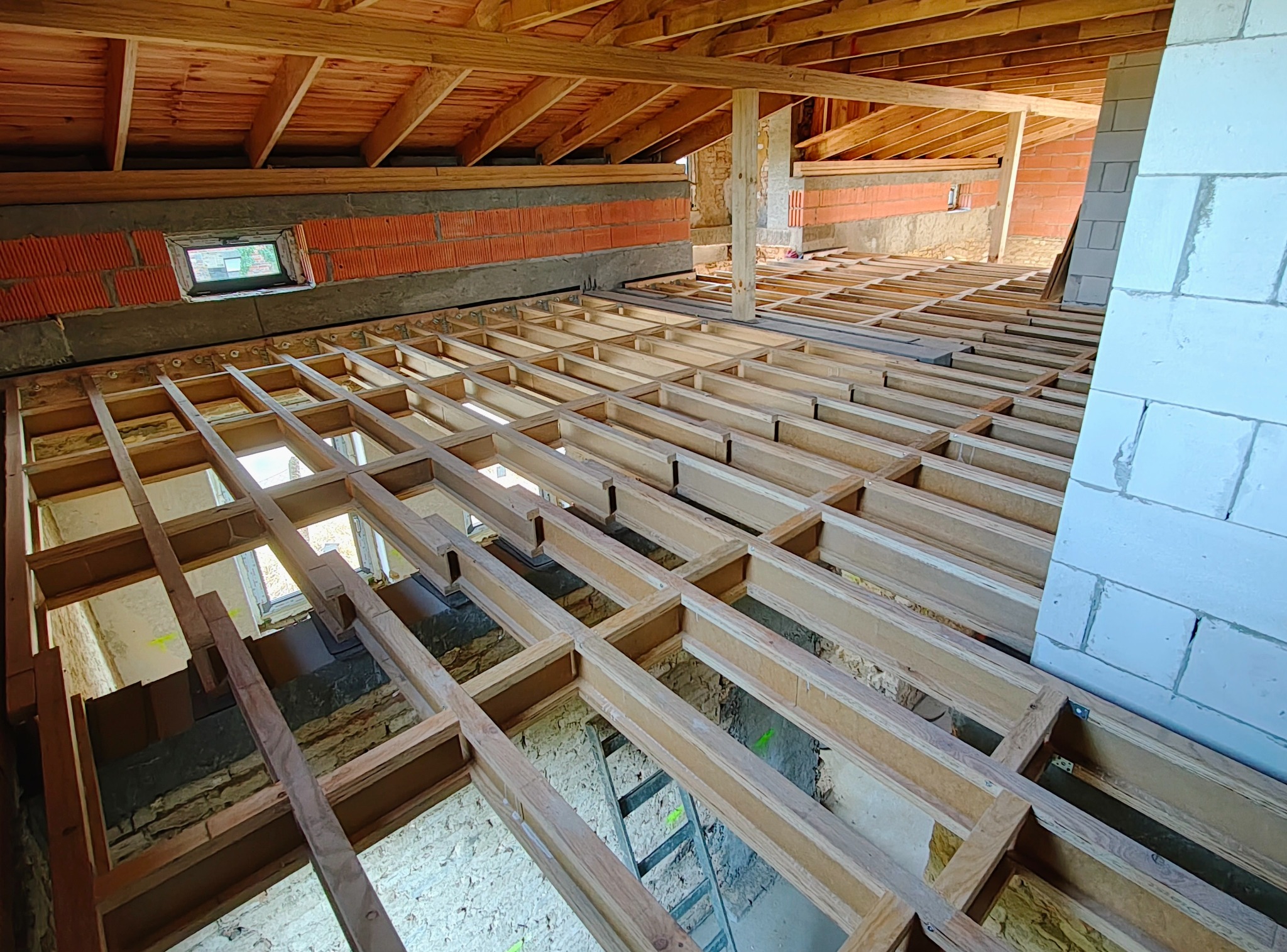 Drewniana konstrukcja podłogi na poddaszu w trakcie budowy, widoczne ceglane ściany i fragment dachu. Ujęcie z góry, z widoczną drabiną prowadzącą na niższy poziom.
