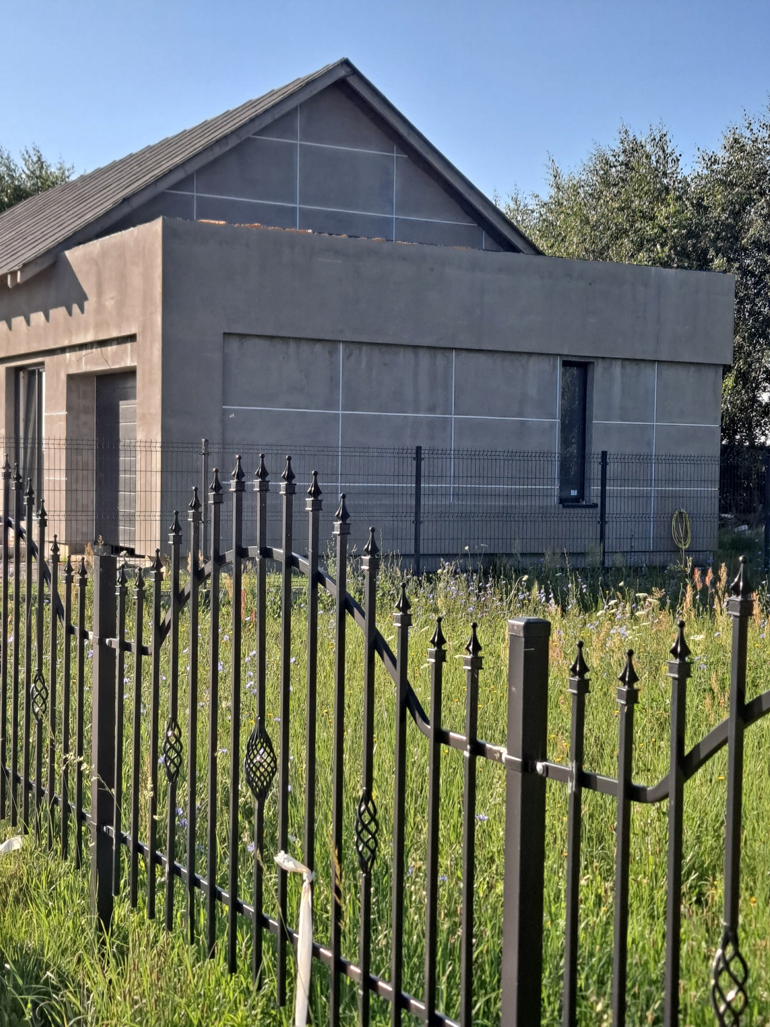 Surowy dom jednorodzinny z szarą elewacją w trakcie budowy, widoczny przez ozdobne, czarne ogrodzenie na tle zielonej trawy i błękitnego nieba.