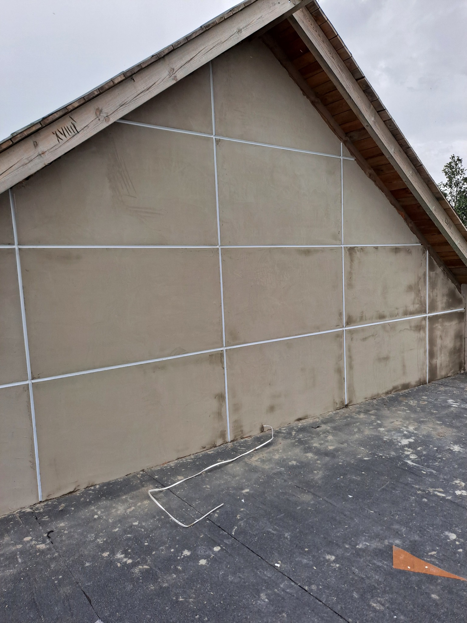 Ocieplona ściana szczytowa budynku z widoczną siatką i nałożonym tynkiem w kolorze beżowym. Widoczne drewniane elementy konstrukcji dachu.